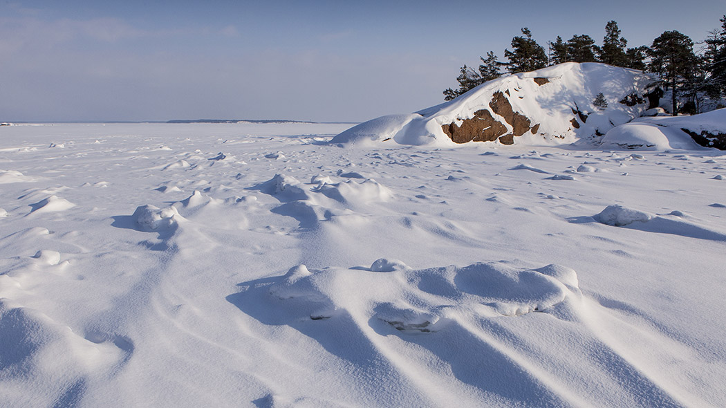 Meri on jäätynyt ja sen päällä on lunta. Jyrkkää kiviluotoa peittää lumi, ja luodon laella kasvaa pieniä mäntyjä.