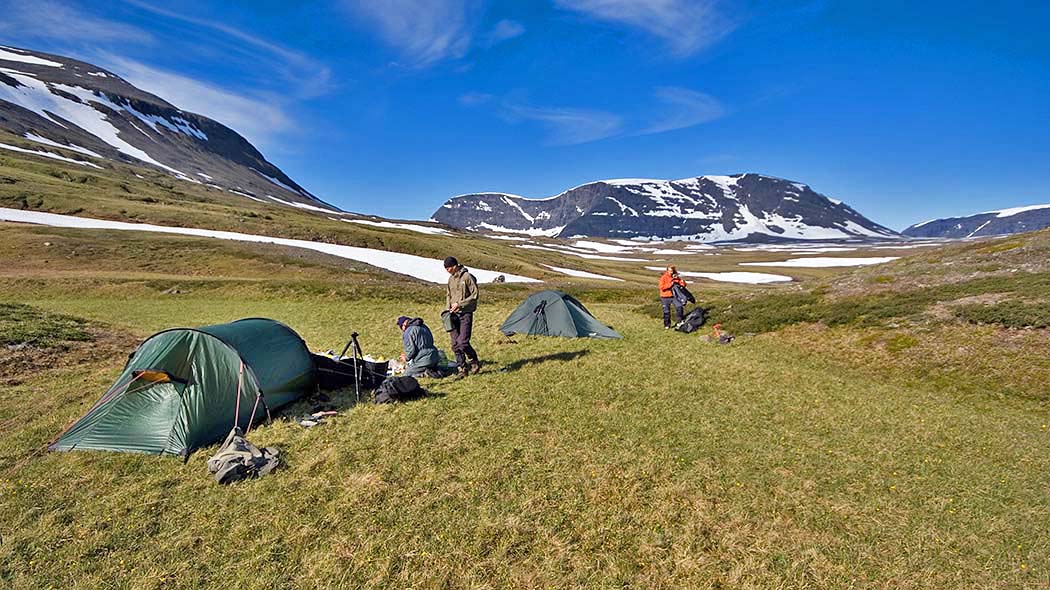 Kolme henkeä pystyttämässä telttoja avoimessa tunturimaastossa aurinkoisessa säässä. Taustalla korkeat tunturihuiput.