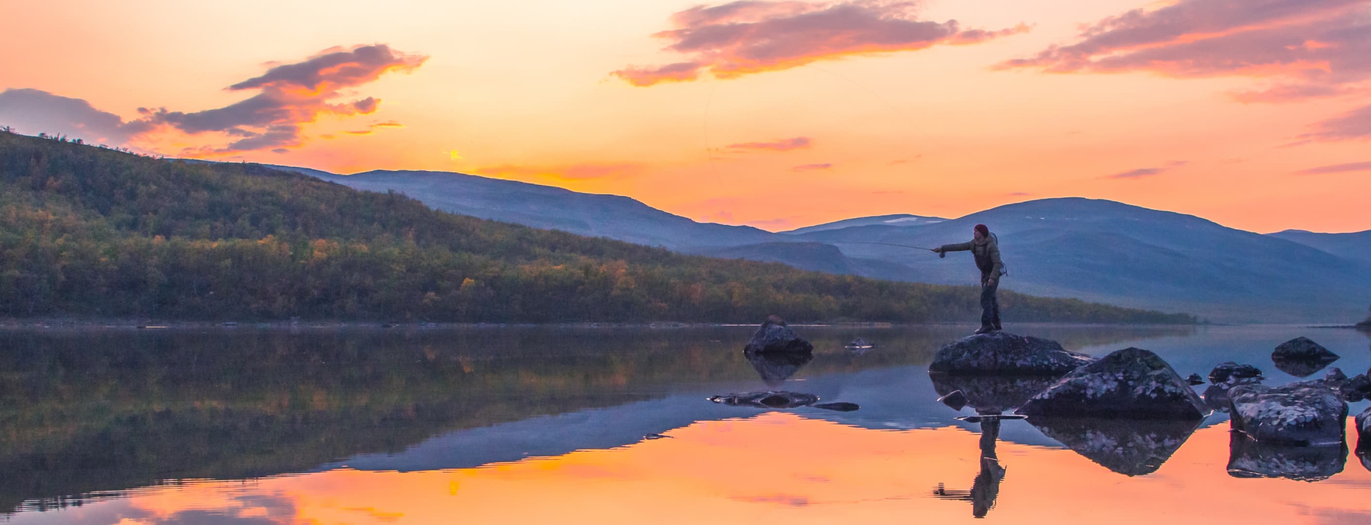 A fisherman stands on a rock in an autumn wilderness. In the background, there are fells, a sunset, and a calm water surface.