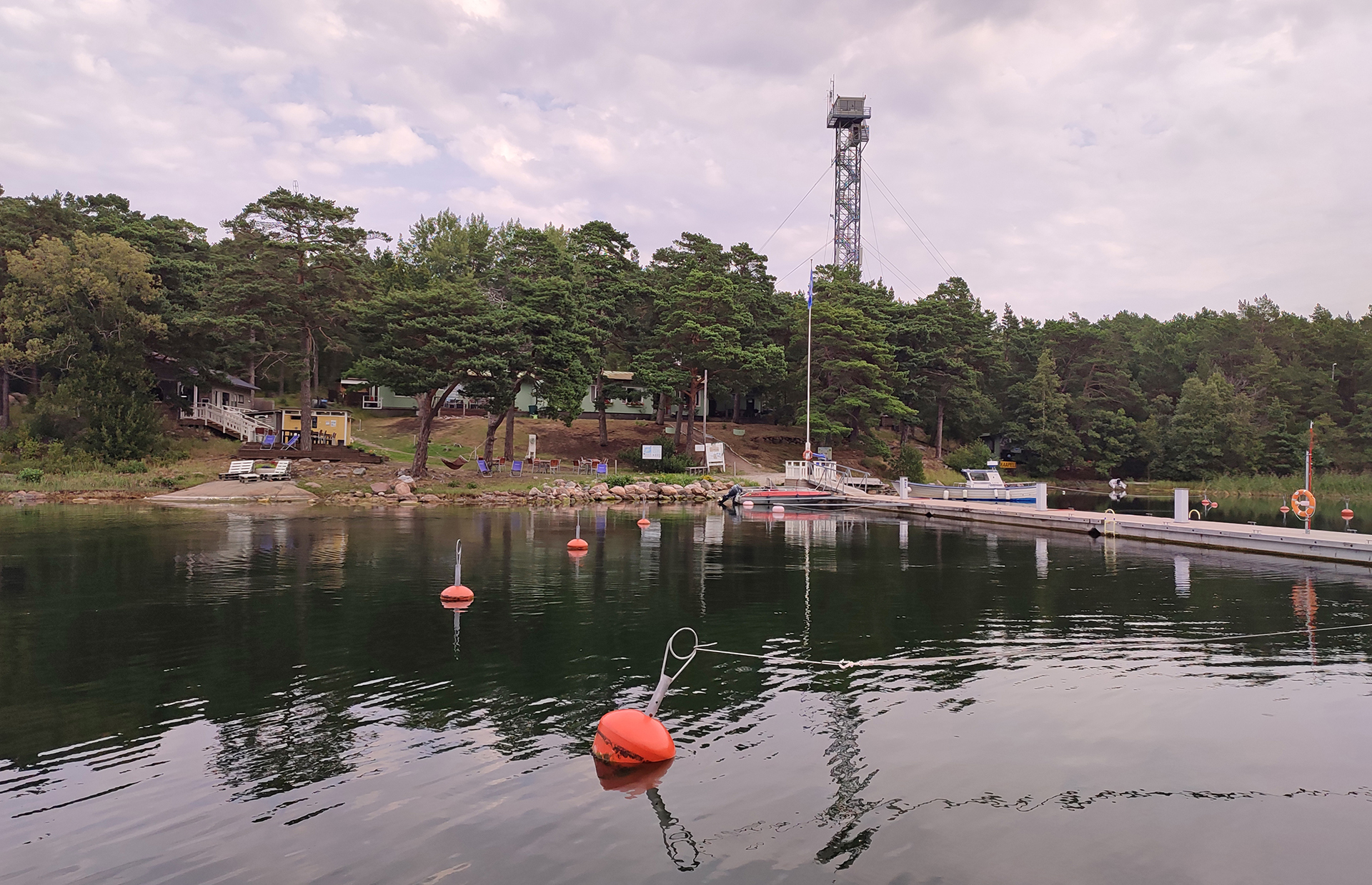 Tyynellä merenlahdella satamassa poijuja, laituri ja rantaa, jossa rakennuksia, metsää ja taaempana korkea torni.