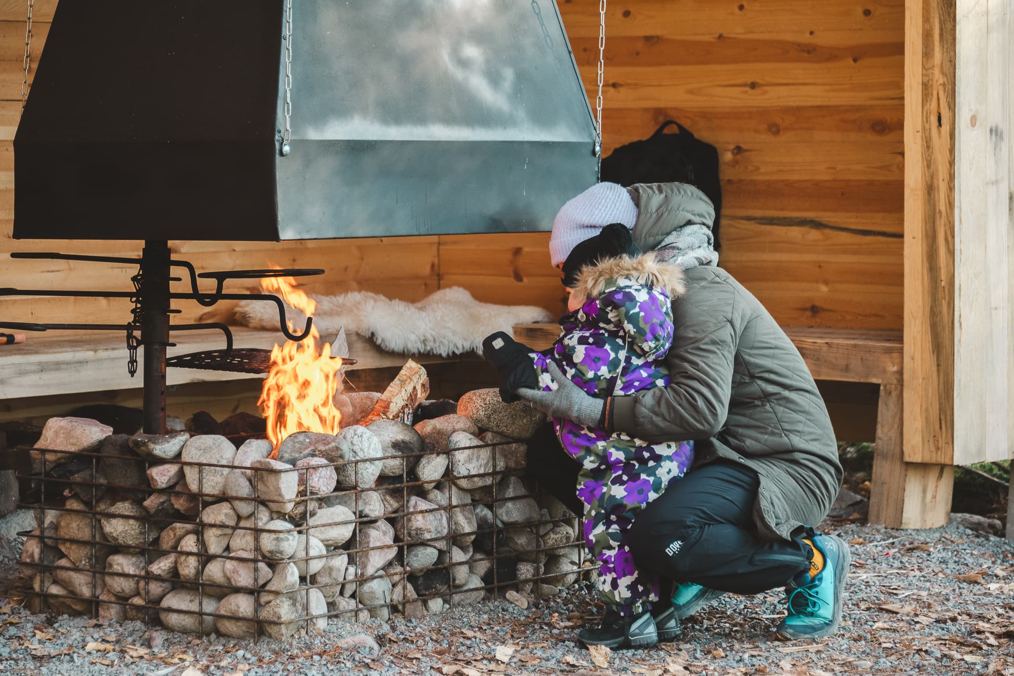 Pieni lapsi ja aikuinen keväisellä retkeilyreitin nuotiopaikalla. Aikuinen on kyykistynyt juttelemaan lapsen tasolle hänen kanssaan tulen ääreen. Tuli loimuaa ja lämmittää retkeilijöitä.