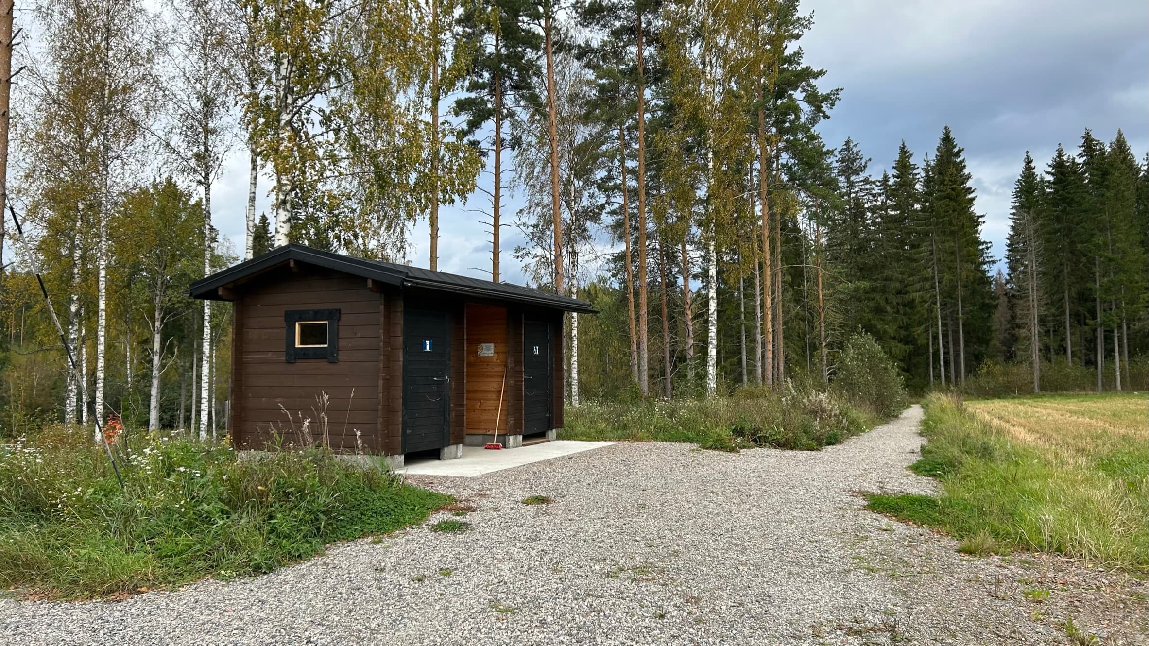 Puurakenteinen ulkokäymälä, jonka luota jatkuu polku. Vasemmalla harvaa koivu- ja mäntypuustoa, oikealla pellon reunaa.