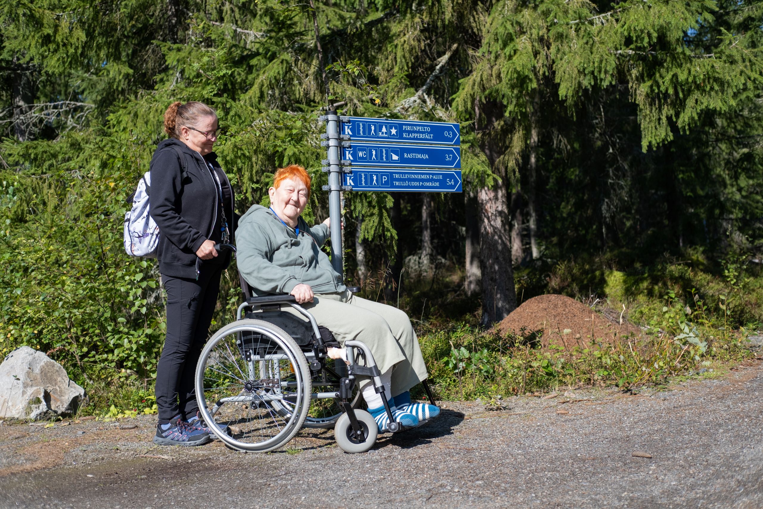 Henkilö istuu pyörätuolissa viittaopasteiden vieressä. Hänen takanaan on avustaja. Viittaopasteen vieressä on muurahaispesä ja taustalla vanhoja kuusia.