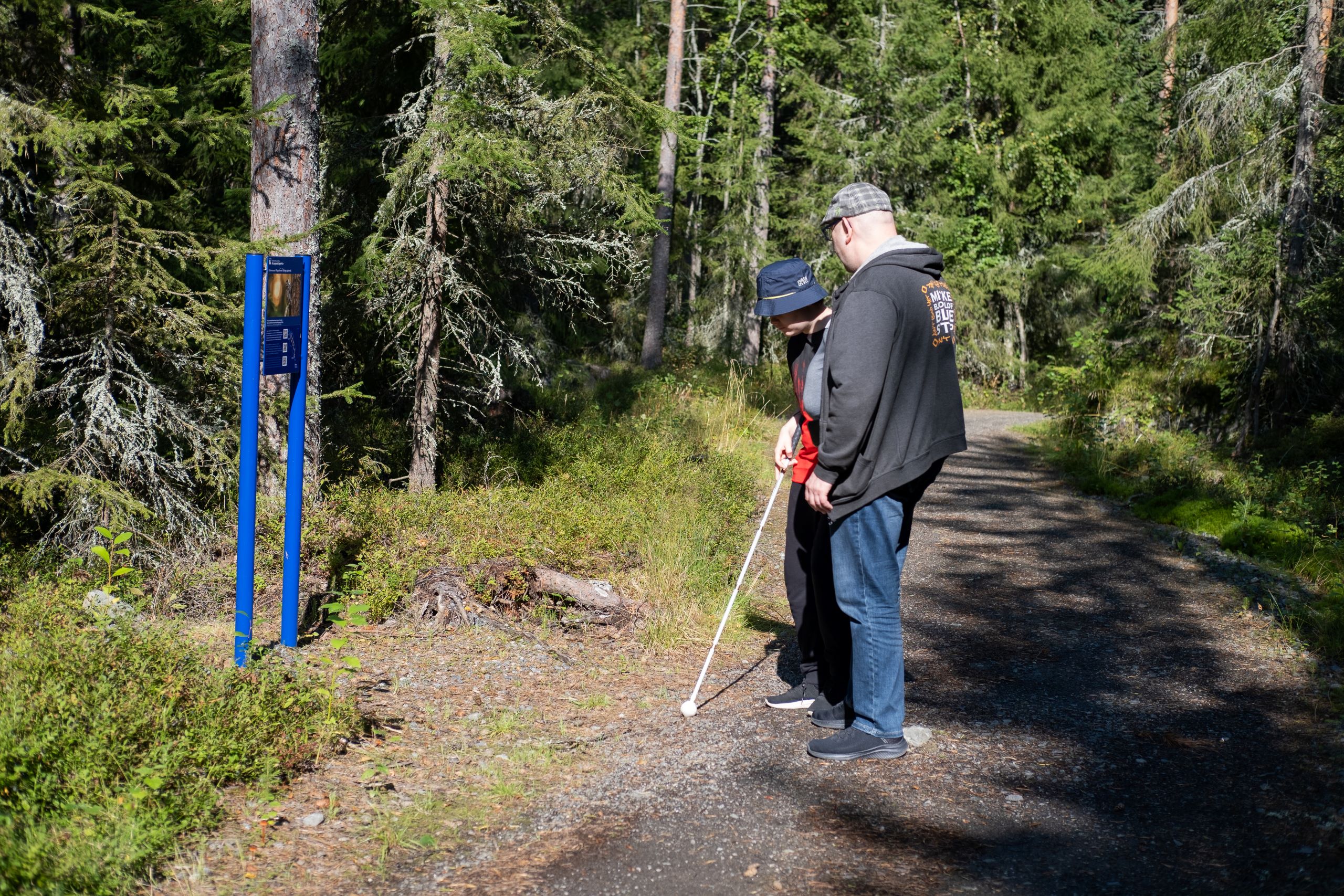 Valkoista keppiä käyttävä henkilö tutustuu avustajansa kanssa polun varren luontopolkuopasteeseen.