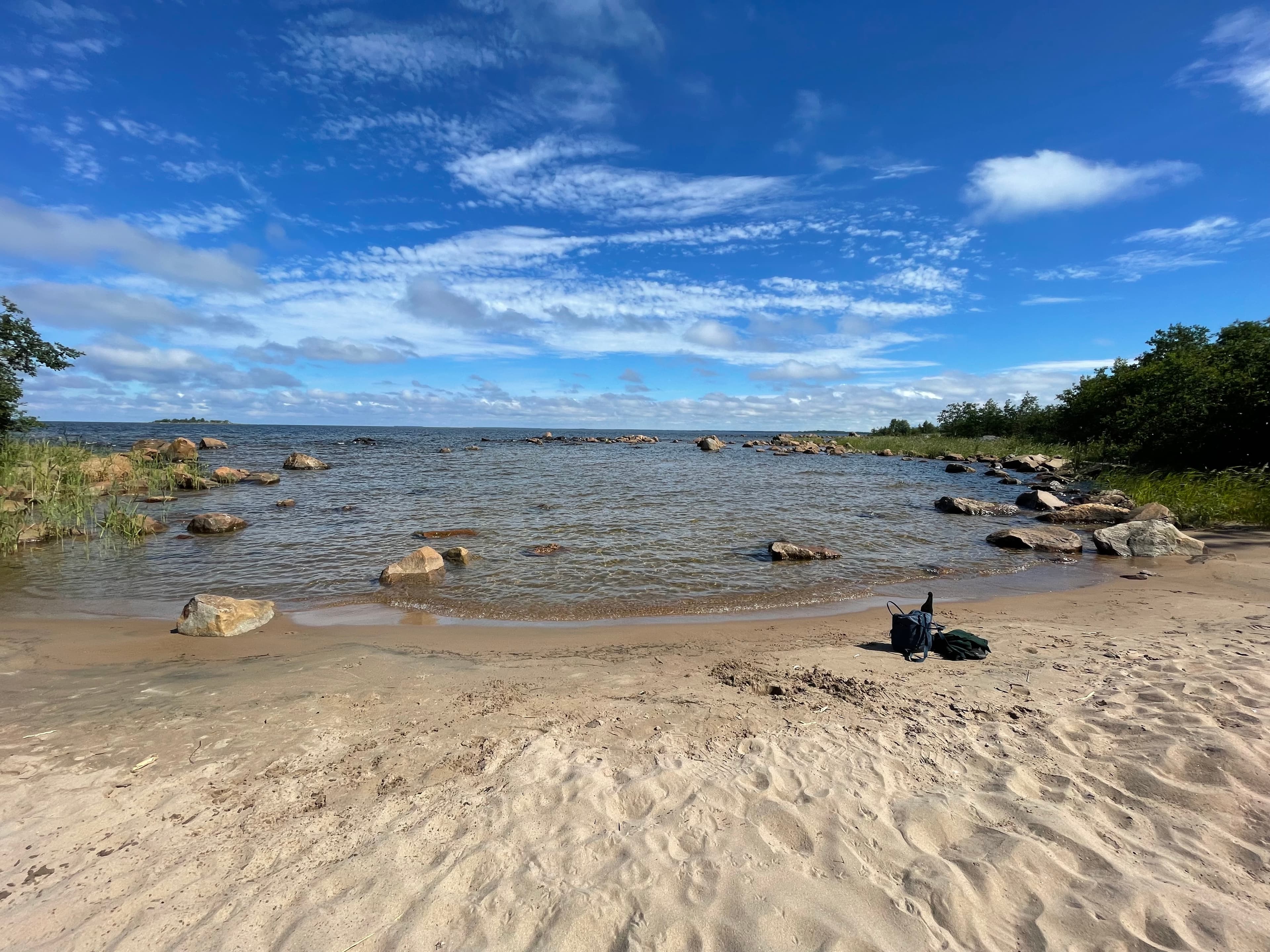 Suojainen hiekkapoukama, jonka reunoilla kiviä ja kasvillisuutta. Taivas on sininen ja taivaalla on hieman valkoisia pilviä. Rannalla on reppu ja paita.