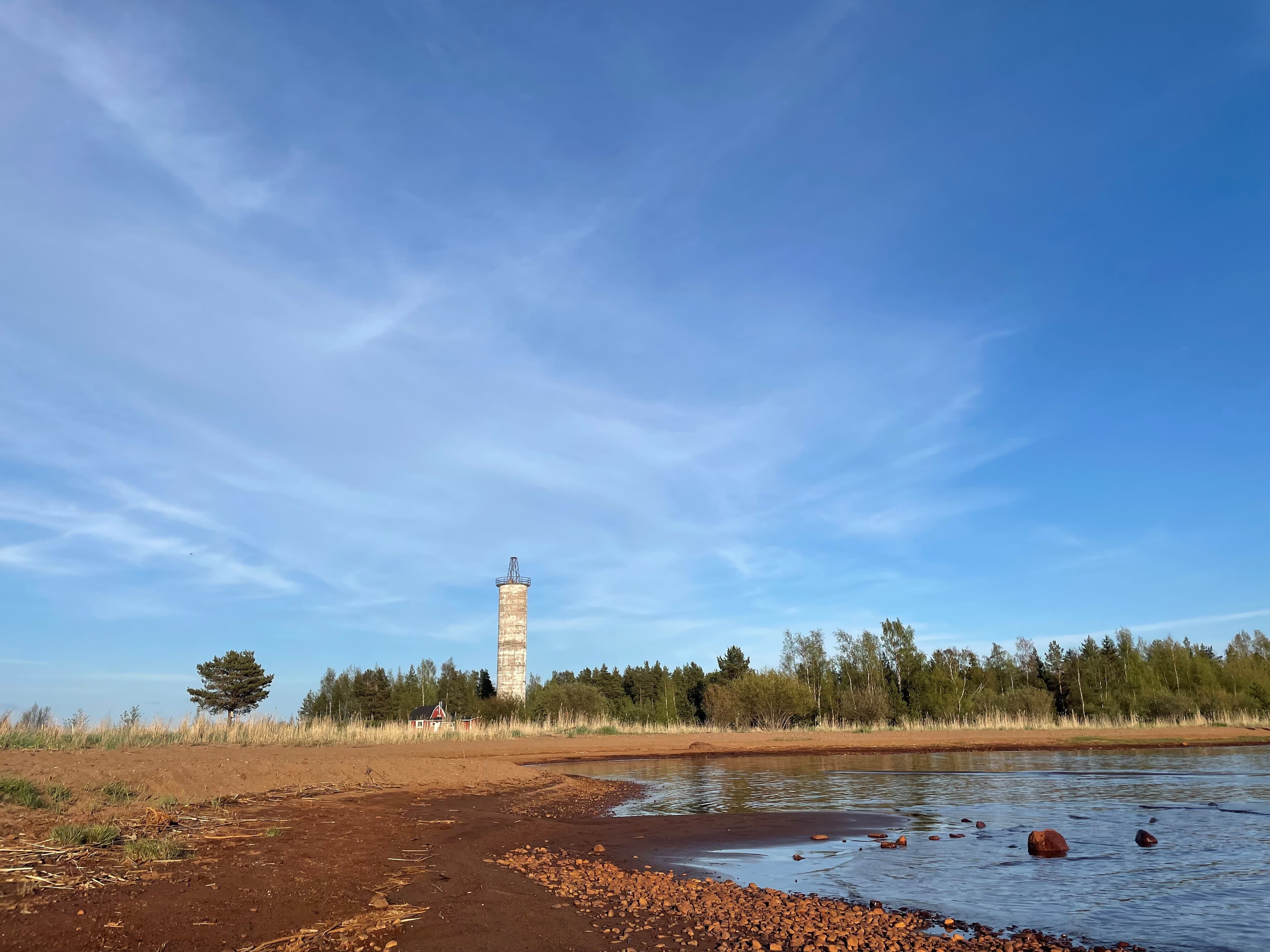 Harrinniemen loisto kuvattuna rannalta päin. Etualallahiekkarantaa ja vettä. Taustalla metsää ja sininen taivas.