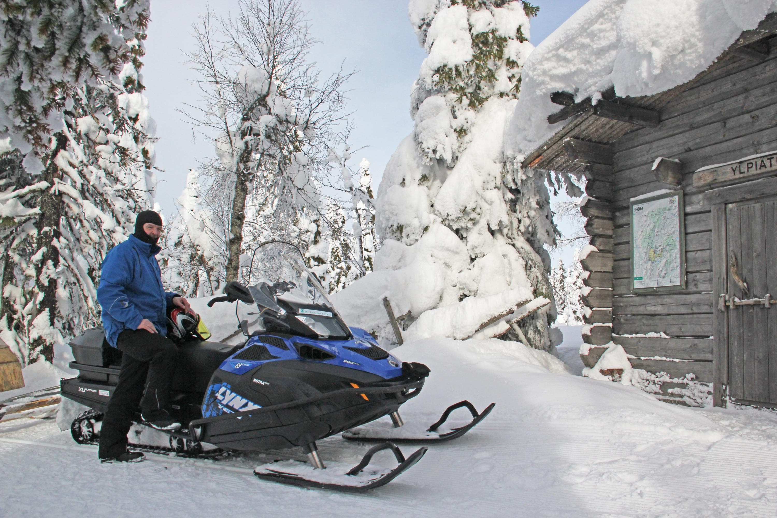 Moottorikelkkailija pysähtyneenä hirsisen tuvan luona.