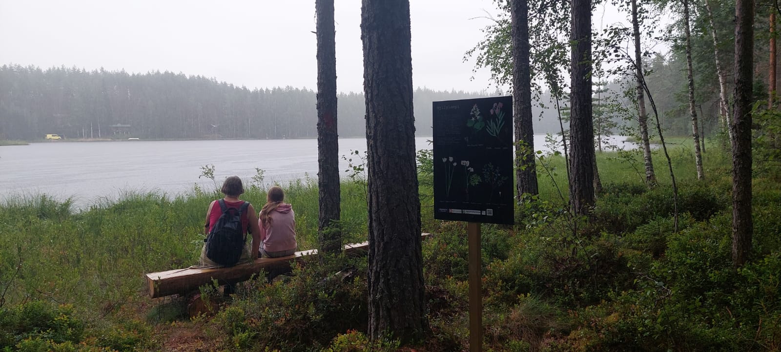 Kaksi naista istuu puupenkillä lammen rannalla ja katsovat lammelle. Etualalla luontopolkutaulu.