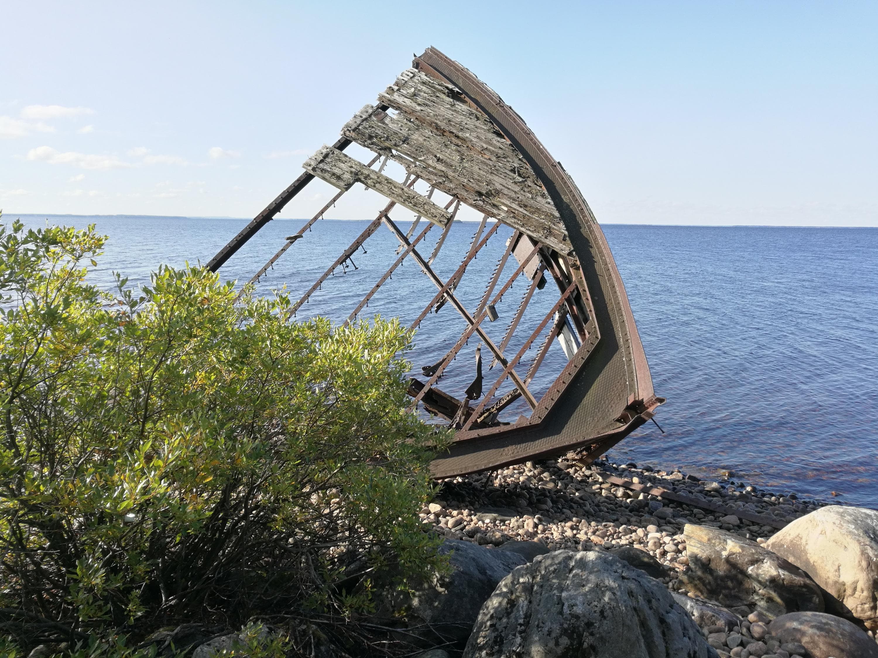 In the middle, the wreck of a barge, in front low vegetation and rocks. In the background, a lake and the sky