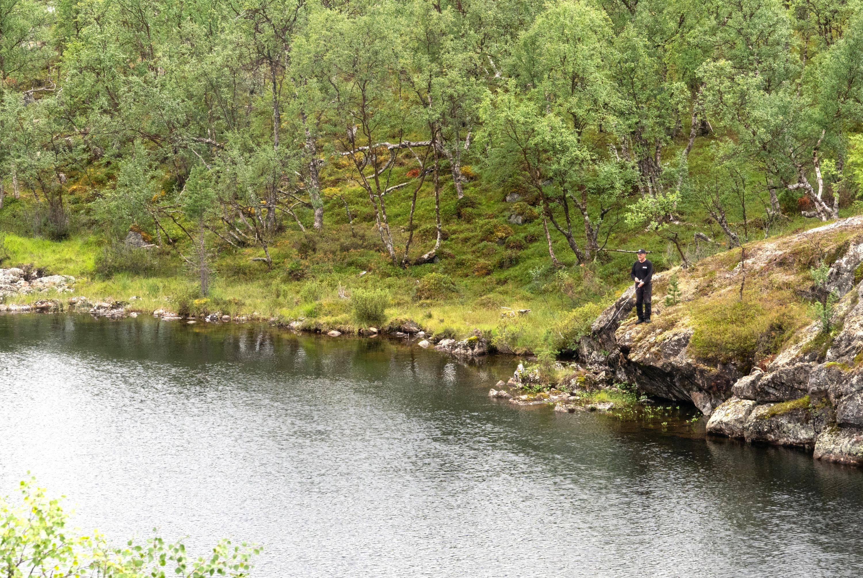 Etualalla vettä, jonka takana kalastaja seisoo jyrkällä kalliolla virveli kädessään. Taustalla koivikkoinen rinne.