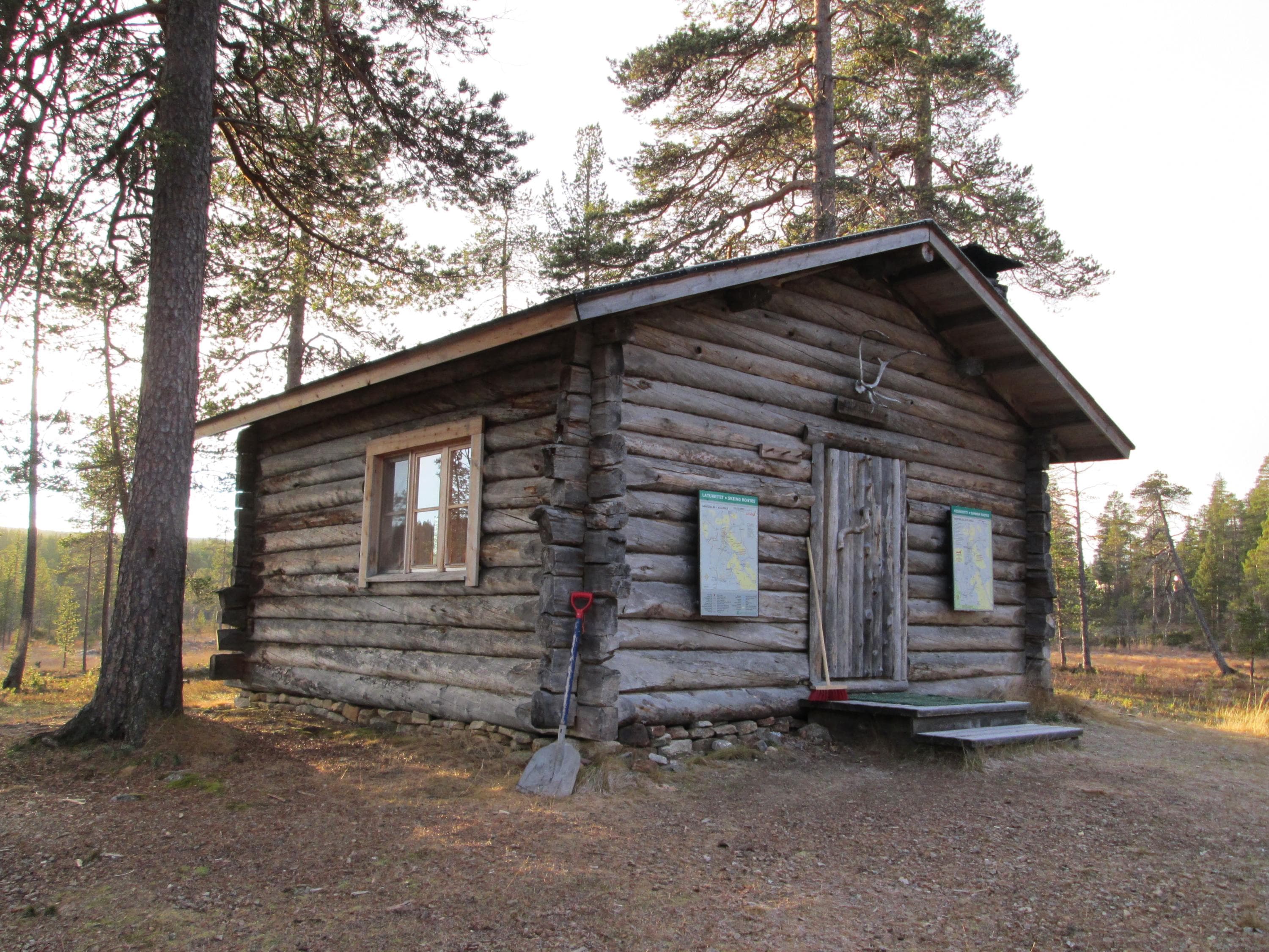 Erämainen tupa, jonka vieressä kasvaa muutama kookas puu. Aurinko paistaa tuvalle. Oven yläpuolella on poronsarvi.