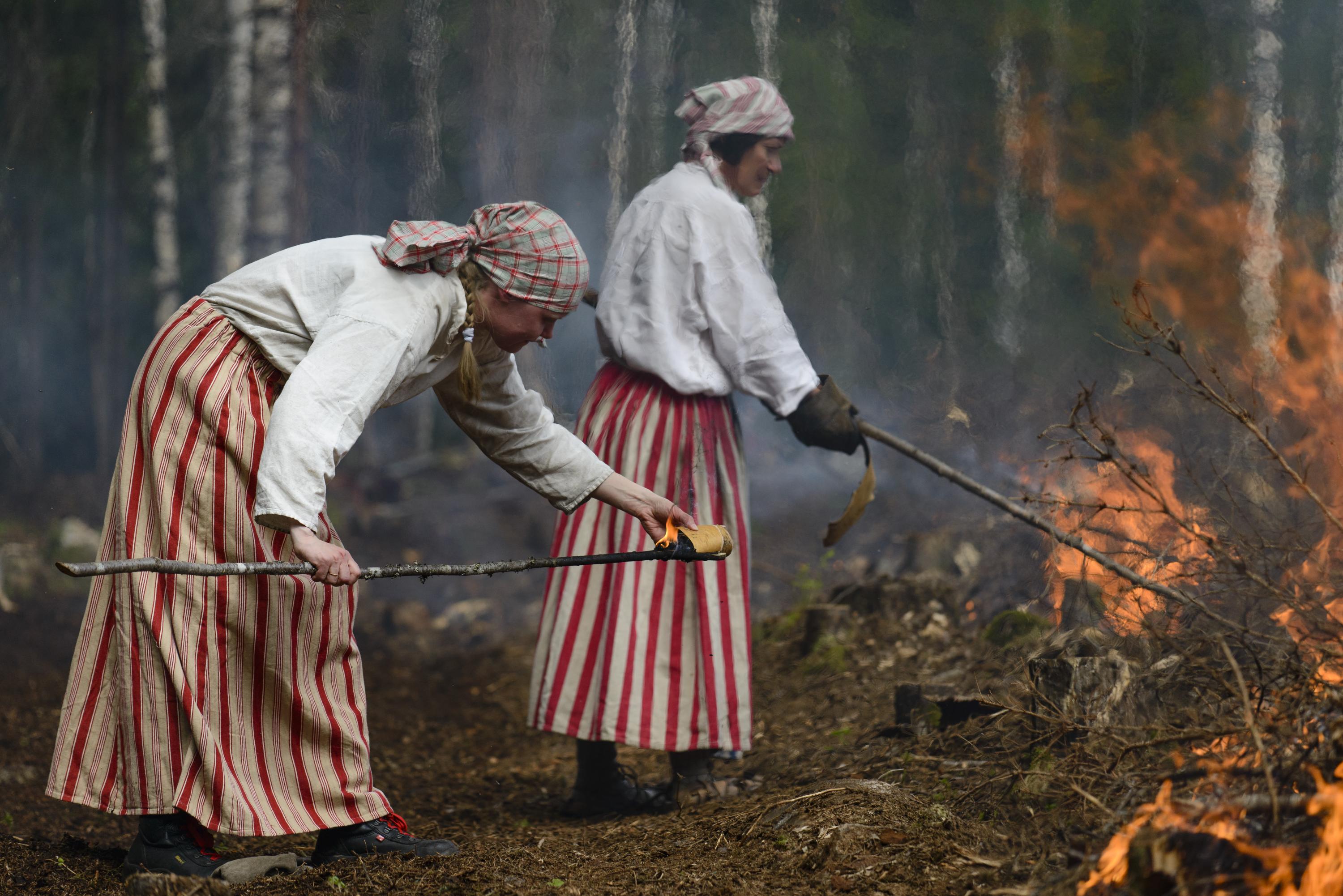 Kaksi punaraidallisiin hameisiin pukeutunutta henkilöä sytyttämässä kaskea. 