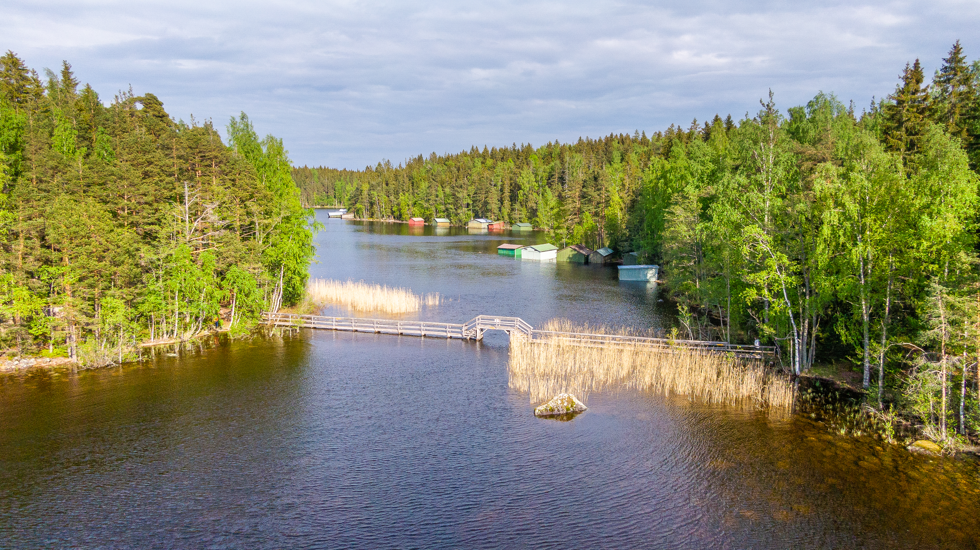 Maisemakuva järvestä, jota ympäröi tiheä metsä. Järven yli kulkee puinen silta ja rannalla on useita venevajoja. Taivas on pilvinen ja vesi heijastaa puiden vihreyttä.