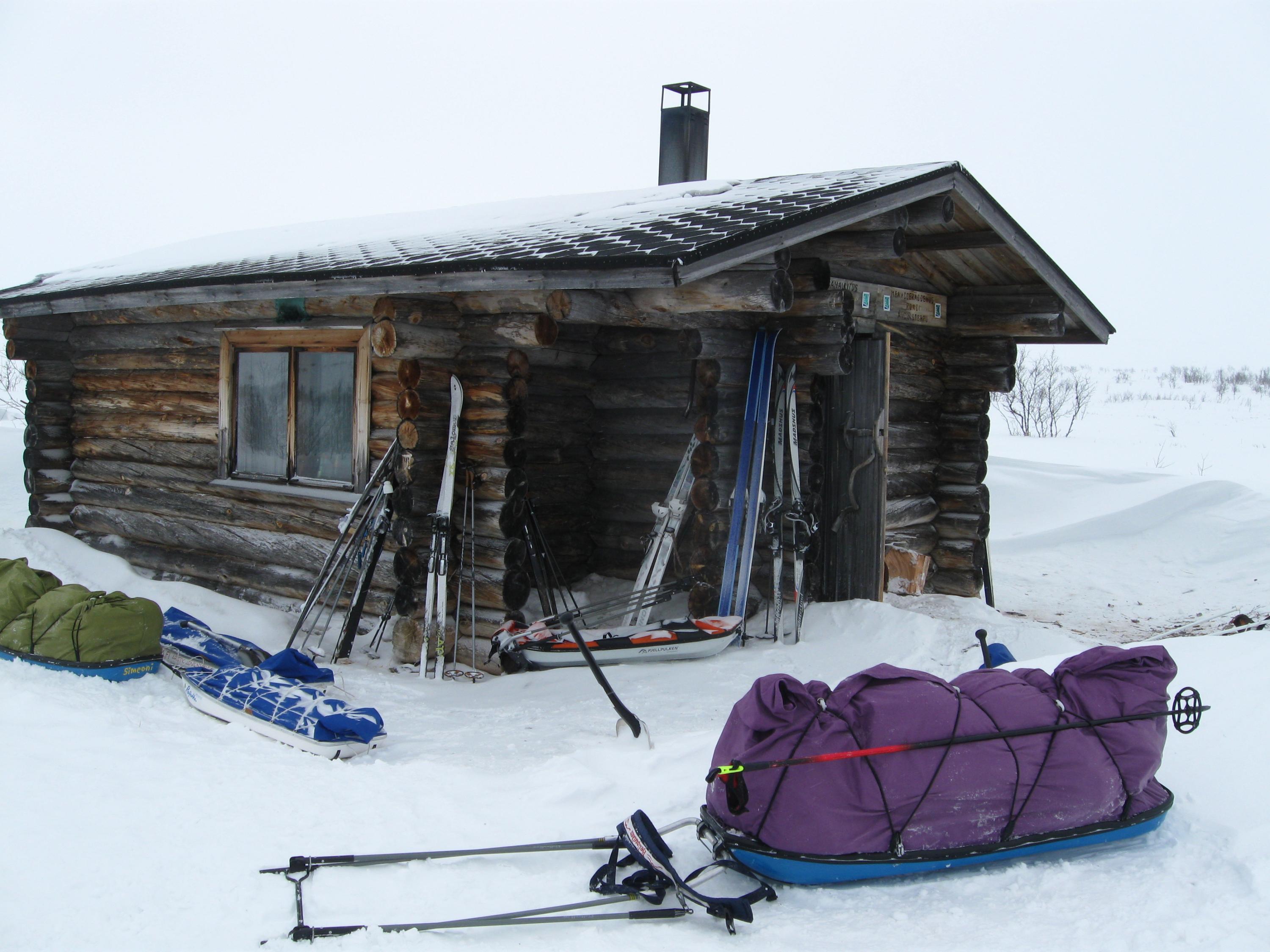 En stuga på vintern. Framför stugan finns flera fjellpulkar och skidor.