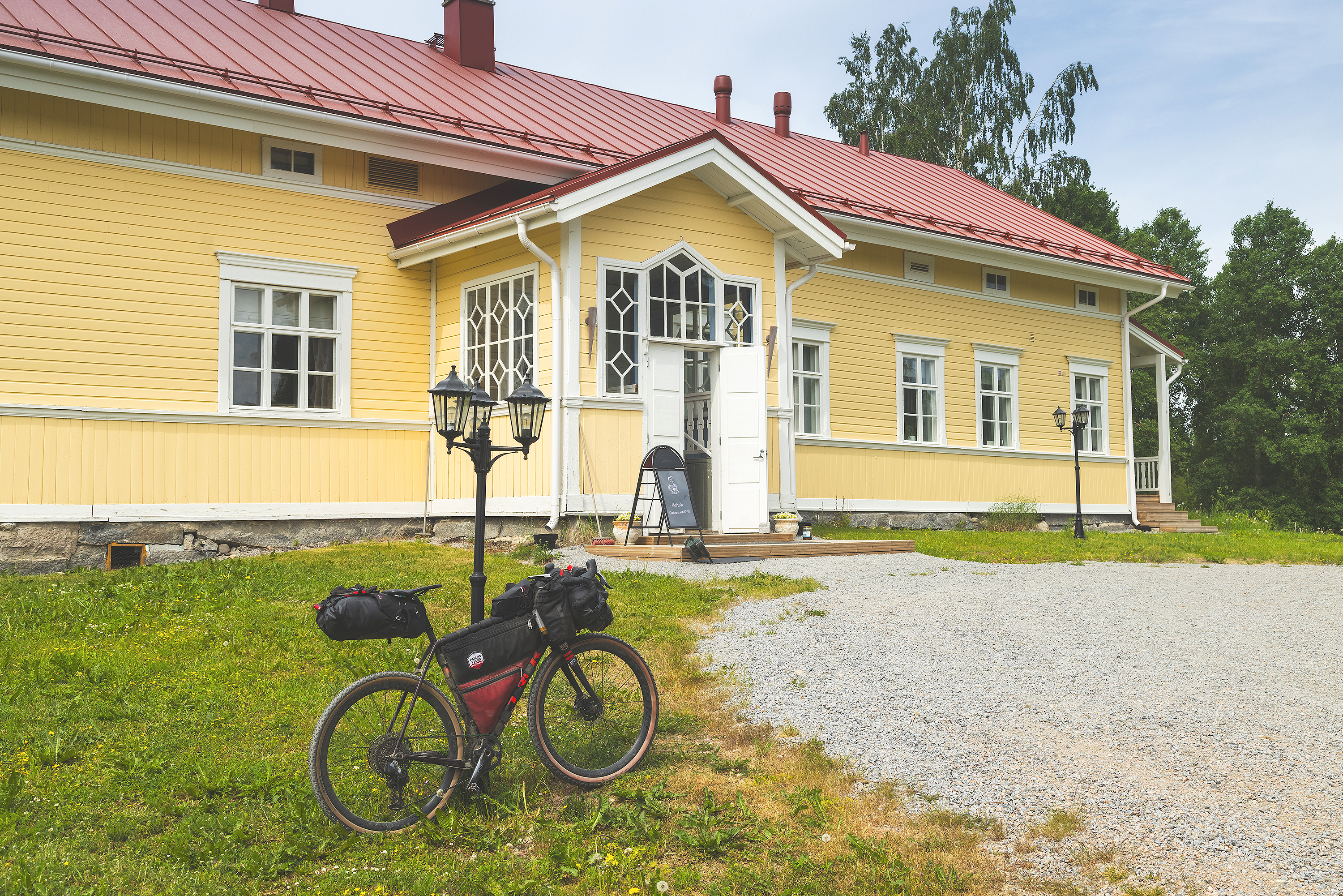 Keltainen maalaisrakennus kesäisessä maisemassa. Rakennuksen edustalla gravel pyörä nojaa valopylvääseen.
