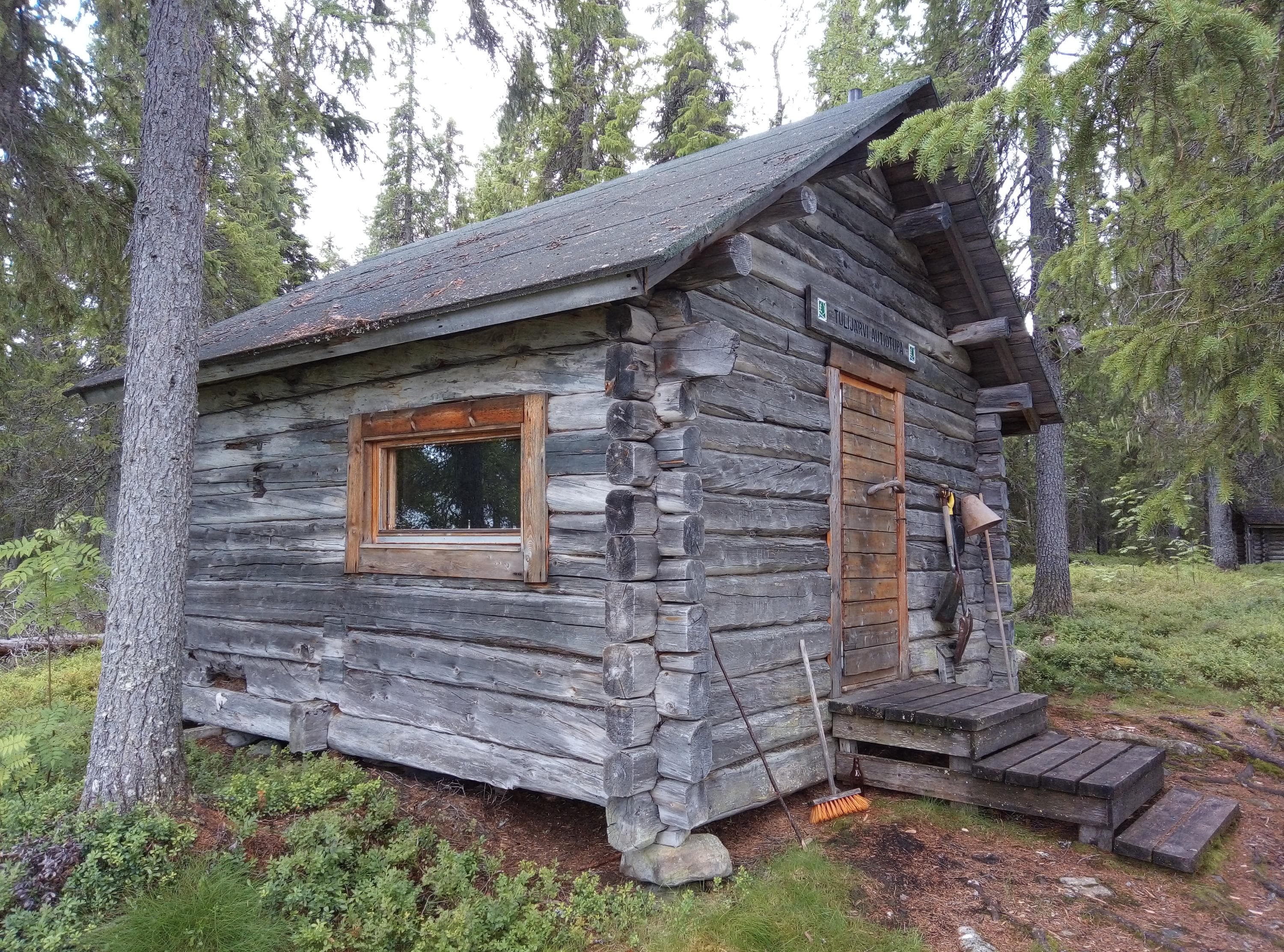 I mitten, en anspråkslös stuga med ett litet fönster. Till vänster om stugan finns en stor tall. Skog i bakgrunden.