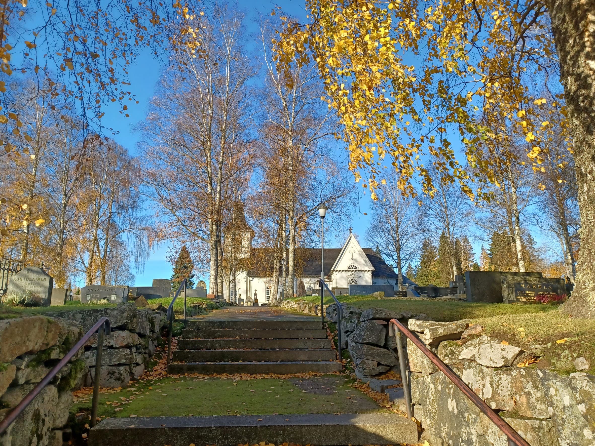 Kuvassa kiviset portaat, jotka johtavat hautausmaalle ja valkoiselle kirkolle. Kuva on otettu syksyllä ja kuvassa olevissa lehtipuissa on vähän keltaisia lehtiä.