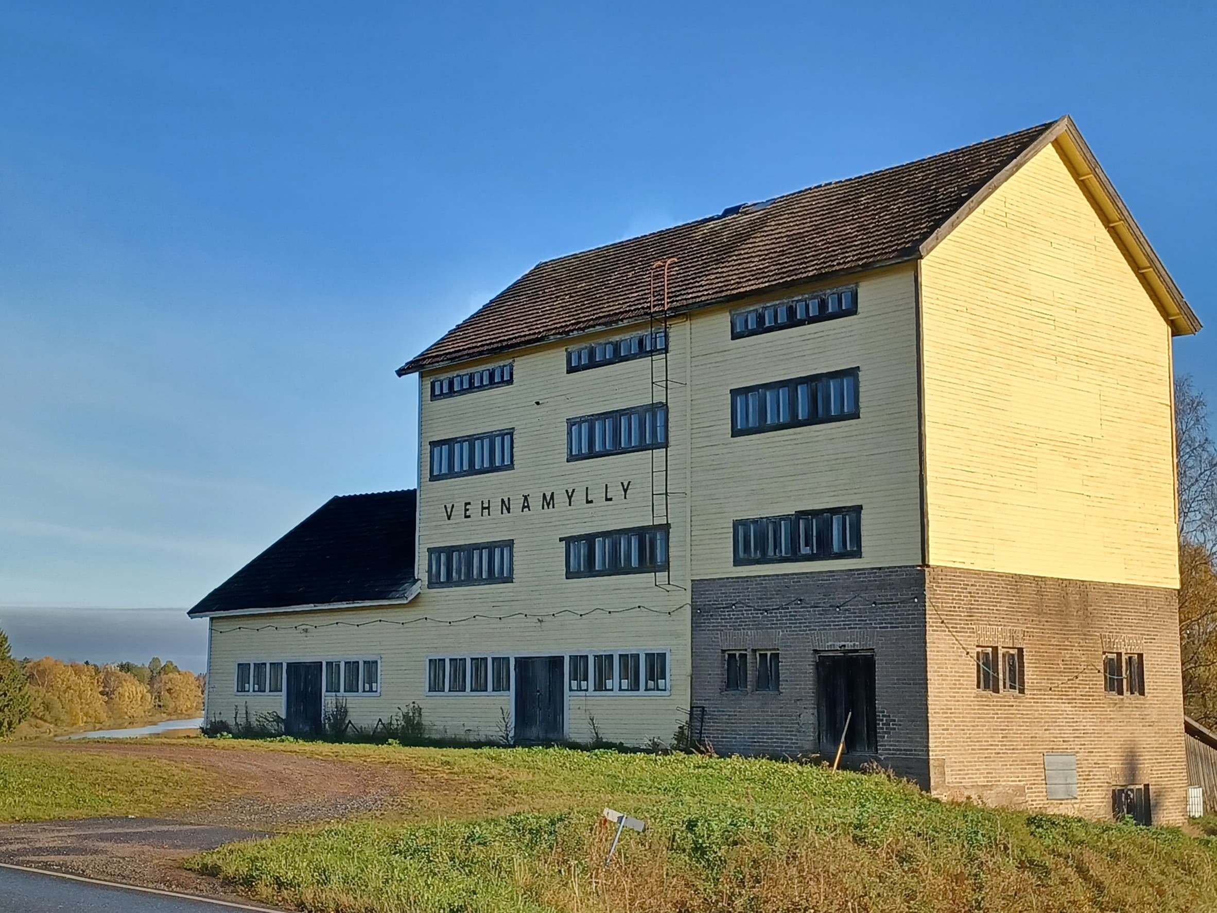 Joen rannassa neljäkerroksinen keltainen myllyrakennus. Rakennuksessa on paljon ikkunoita.