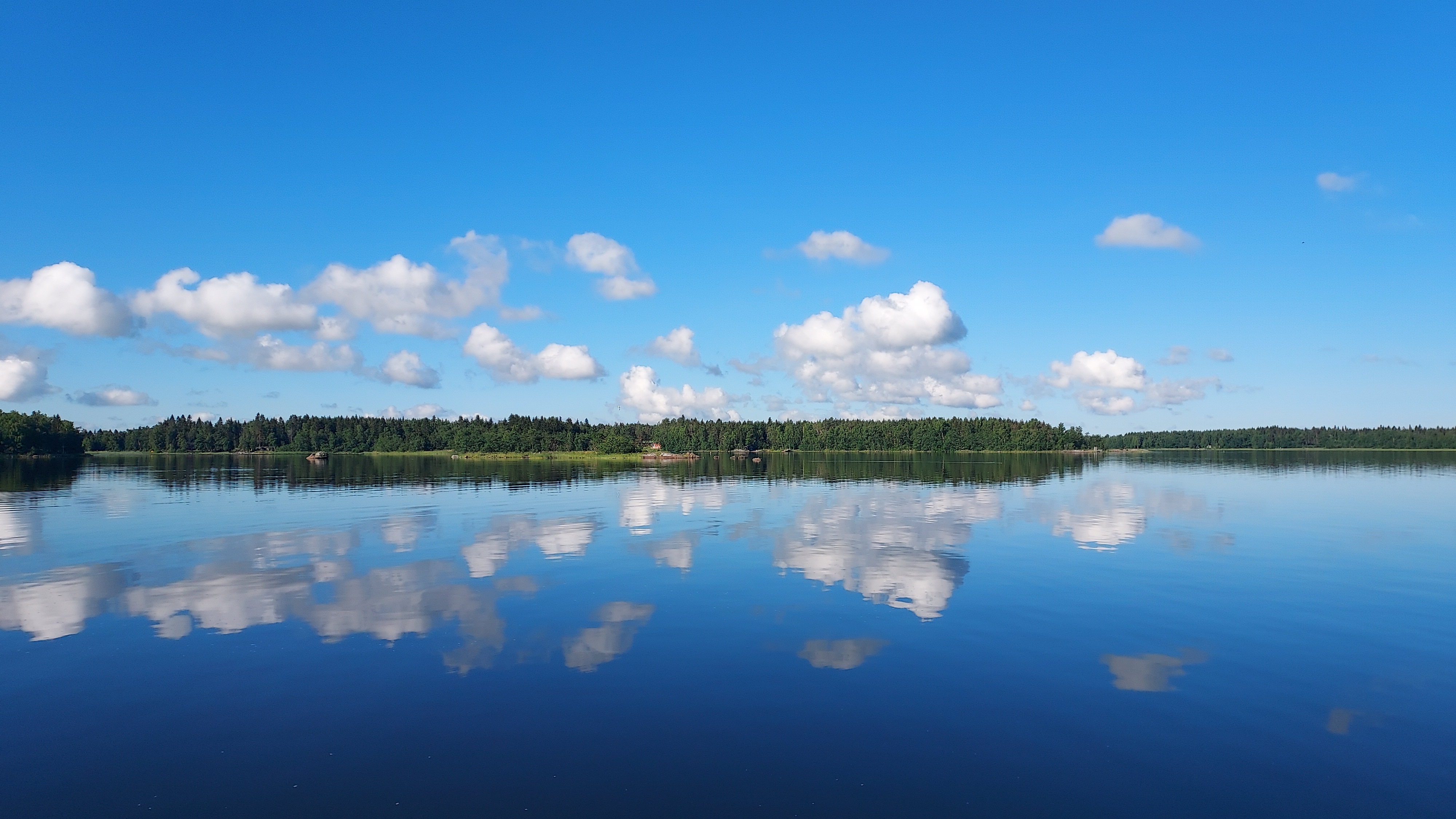 Maisema Isokarin saarelta. Peilityyni meri, jonka pintaan heijastuvat pilvet siniseltä taivaalta sekä vastakkaisen rannan metsä.