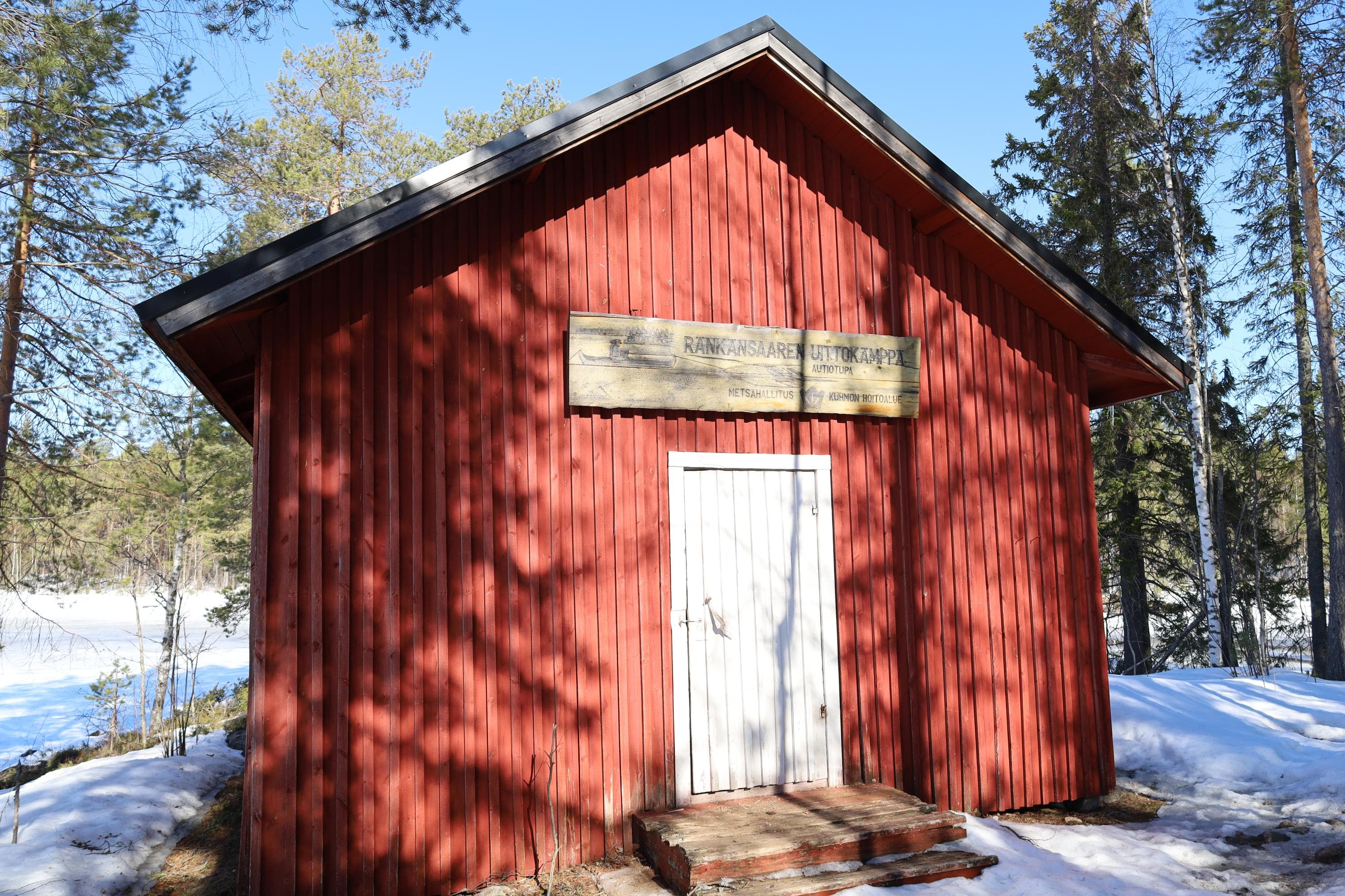 Keskellä lautarakenteinen tupa, taustalla metsää ja taivas. Ympärillä lunta.
