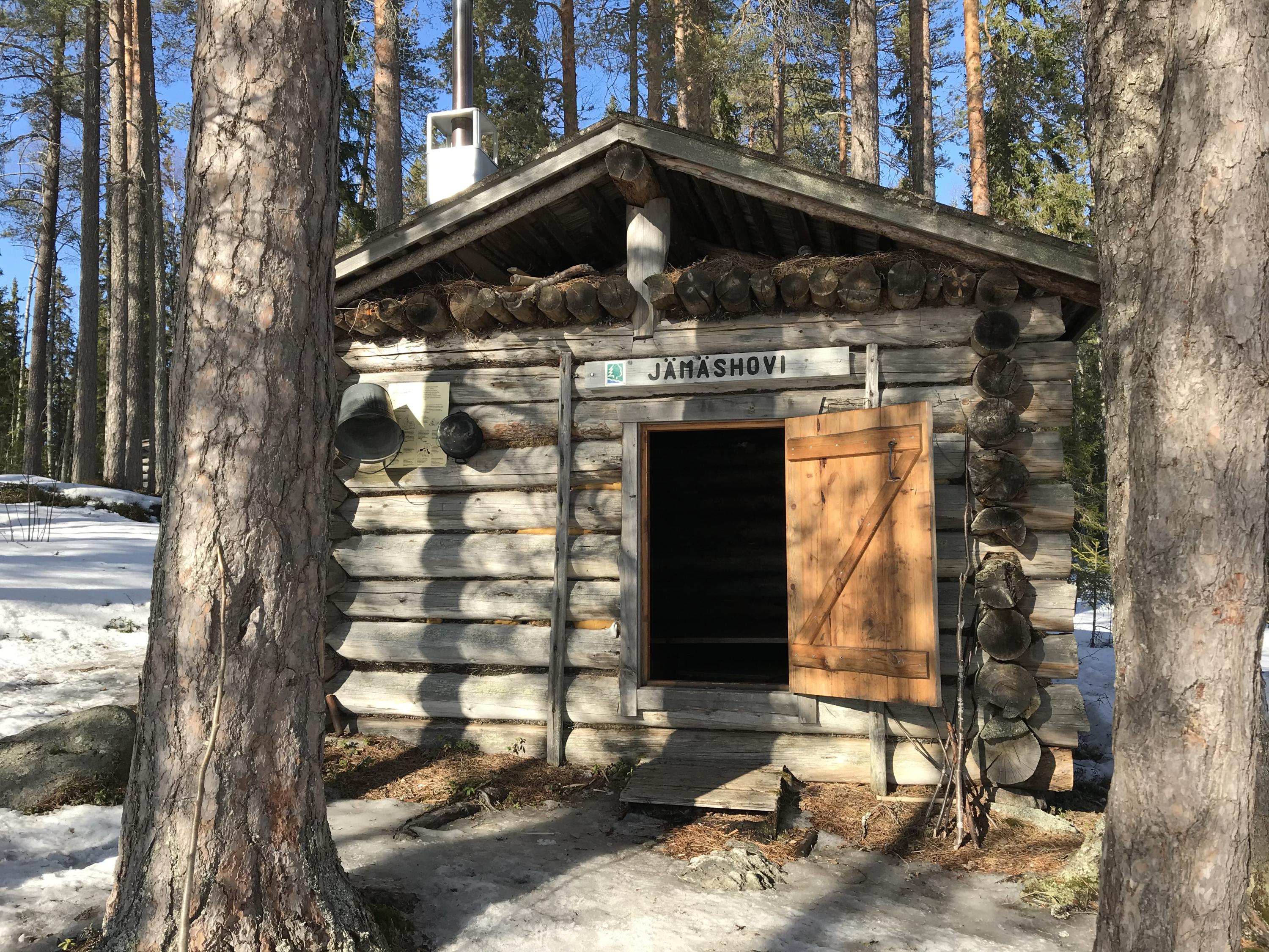 Kuvan keskellä pieni hirsinen rakennus, jossa ovi auki. Vasemmassa ja oikeassa reunassa suuret puunrungot.