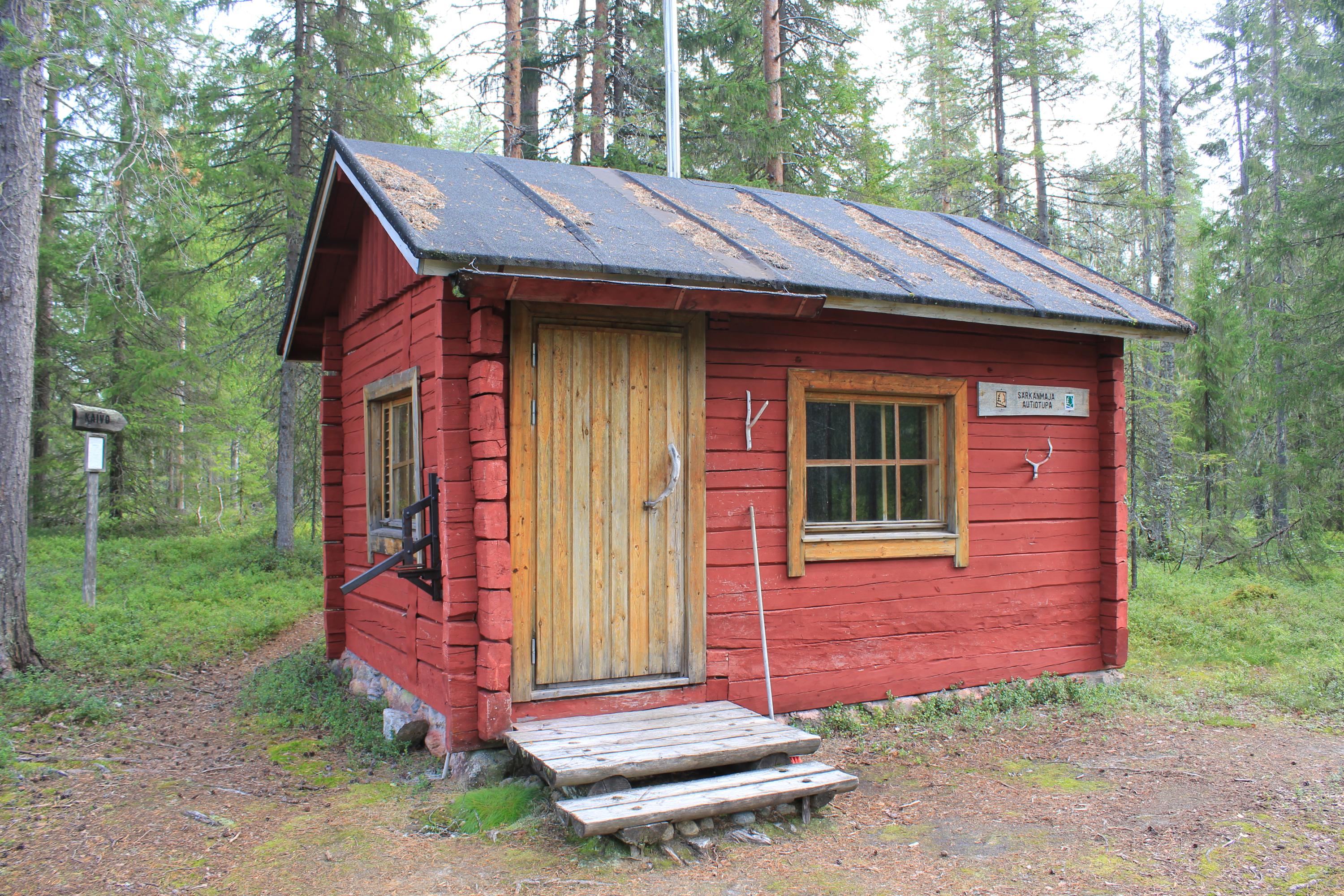 Keskellä kuvassa pieni mökki, jossa ovi vasemmalla, ikkuna oikealla. Taustalla metsää. 