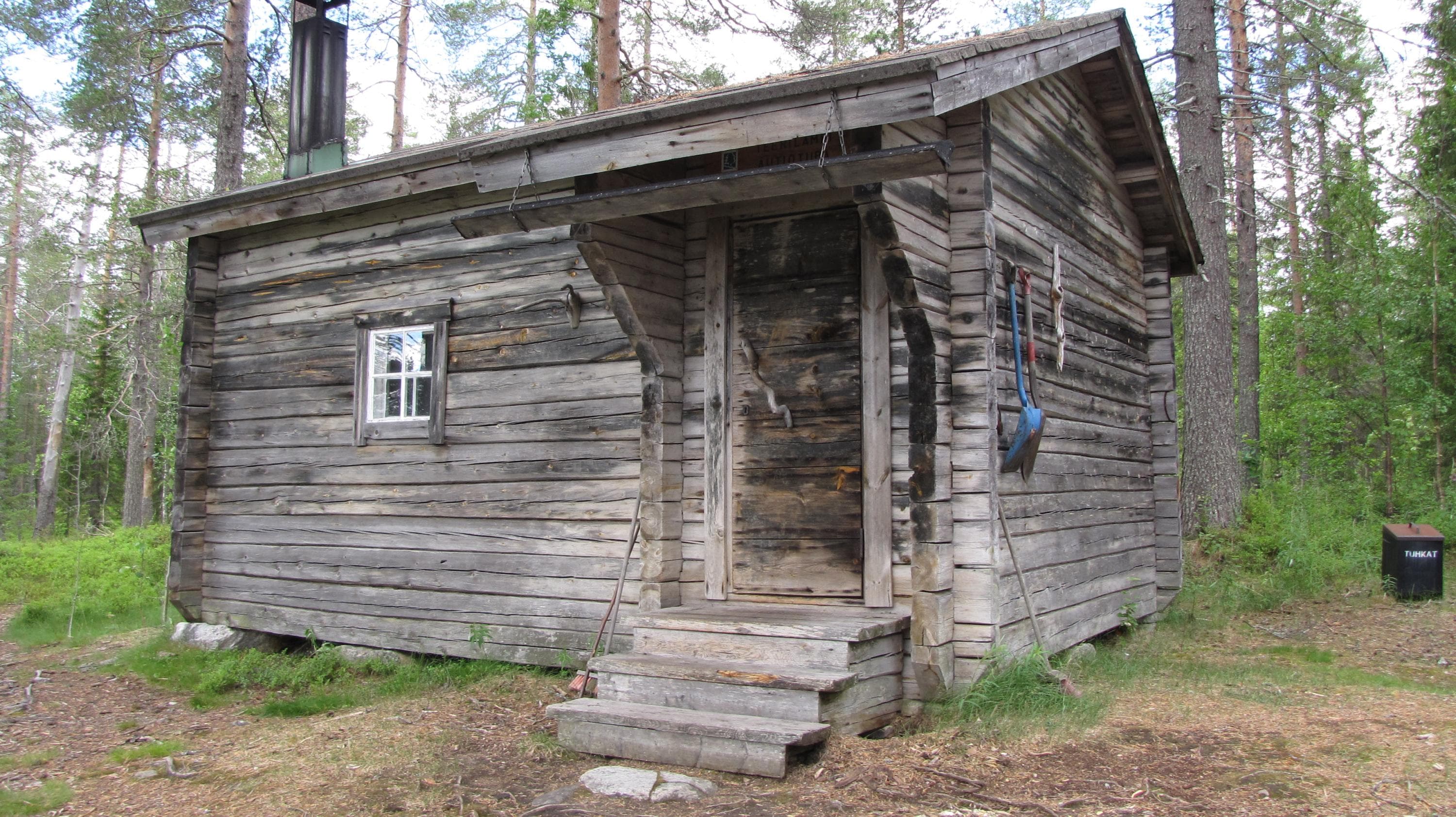 Keskellä tupa, jonka oikeassa reunassa on portaat ja ovi sekä katos. Taustalla metsää. 