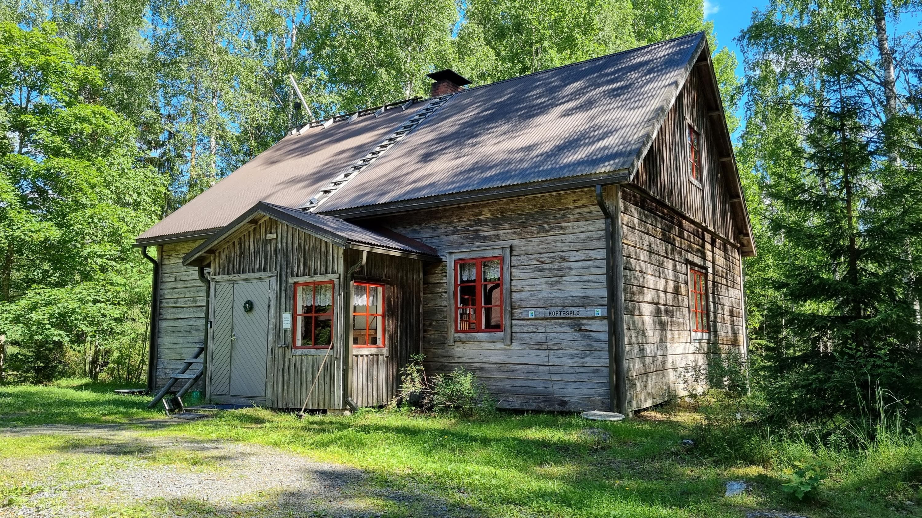 Rustic wooden house with a slanted roof and red-framed windows. Surrounded by green trees and vegetation.