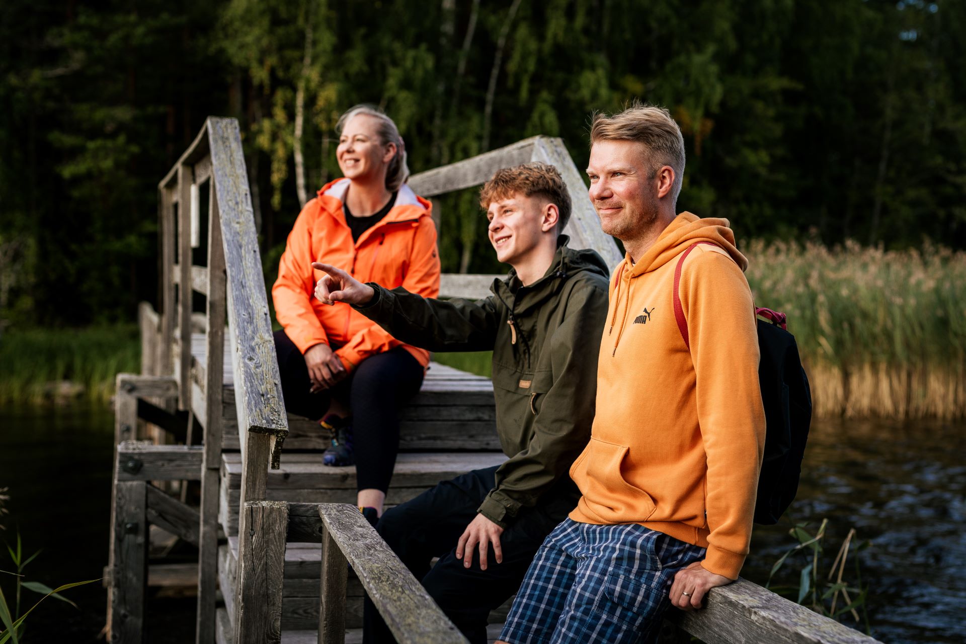 Kolme ihmistä istuu puisella sillalla luonnon keskellä. Yksi henkilö osoittaa eteenpäin, ja taustalla näkyy metsää ja vesistöä.