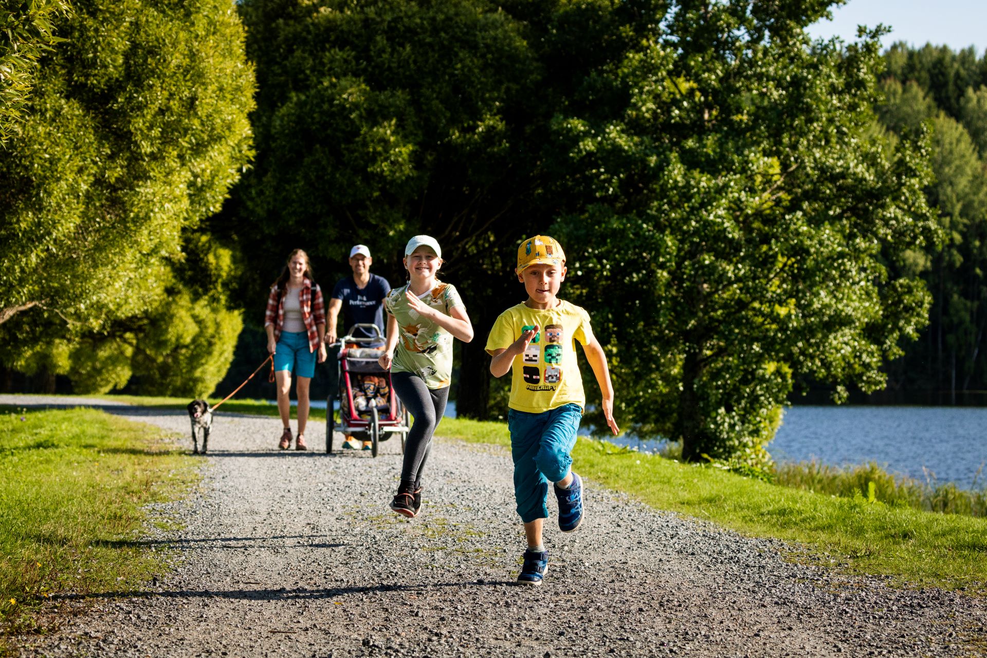 Kuvan etualalla kaksi lasta juoksemassa. Kuvan taka-alalla pariskunta, joista toinen työntää lastenvaunuja ja toinen taluttaa koiraa.