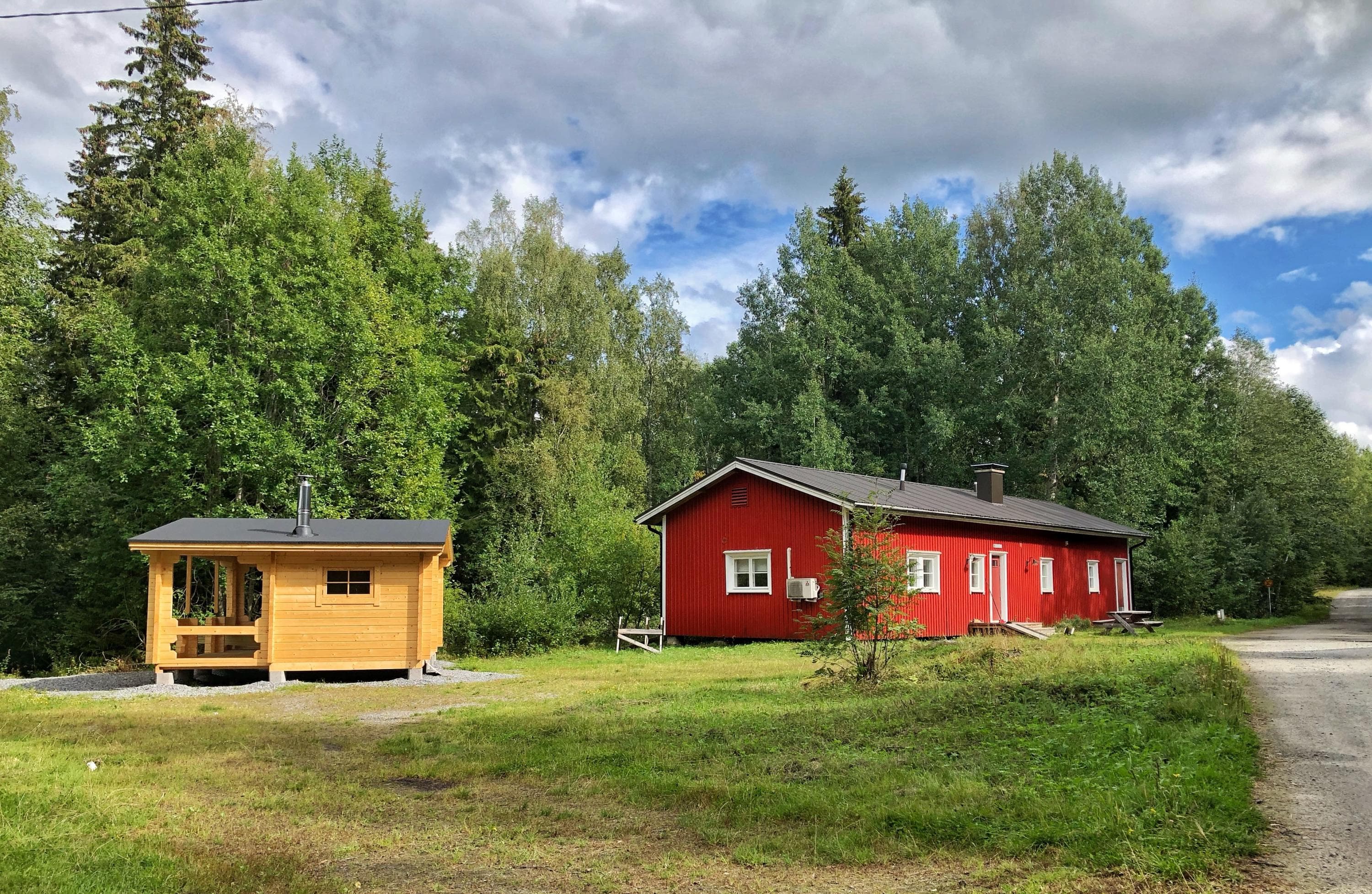 Rural scene with two buildings. On the left, a small wooden structure; on the right, a larger red building. Surrounded by grass and trees.