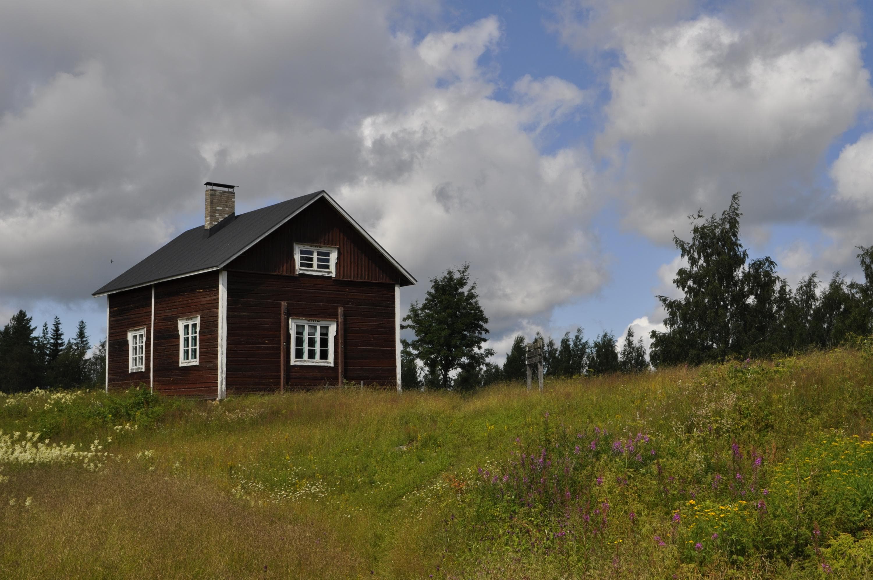 Keskellä vasemmassa reunassa vanha hirsirakennus. Kuvan alaosassa vanhaa peltoa. Yläosassa taivas.