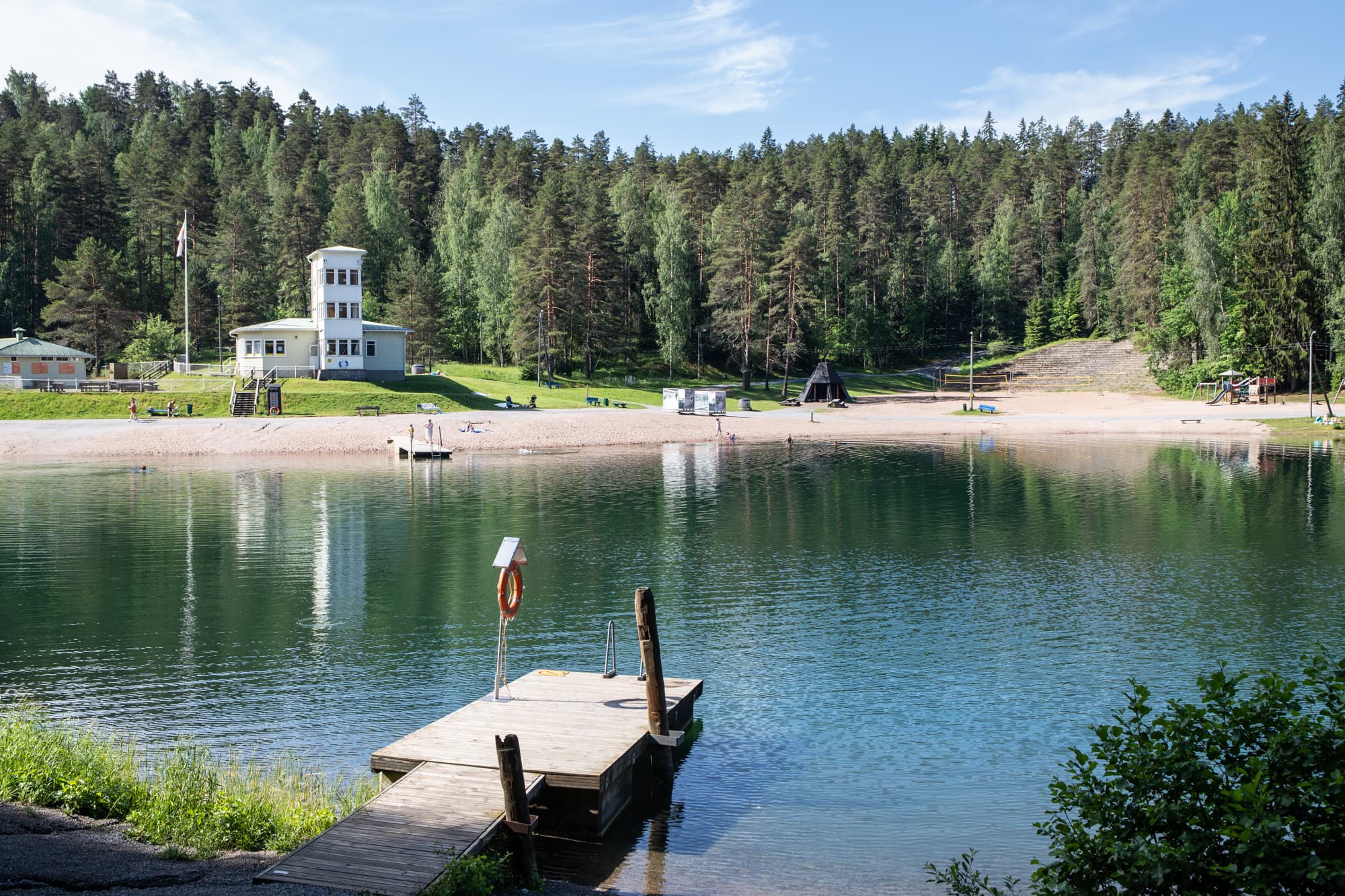 Edessä laituri. Taustalla hiekkaranta, järvi, kahvilarakennus ja metsää