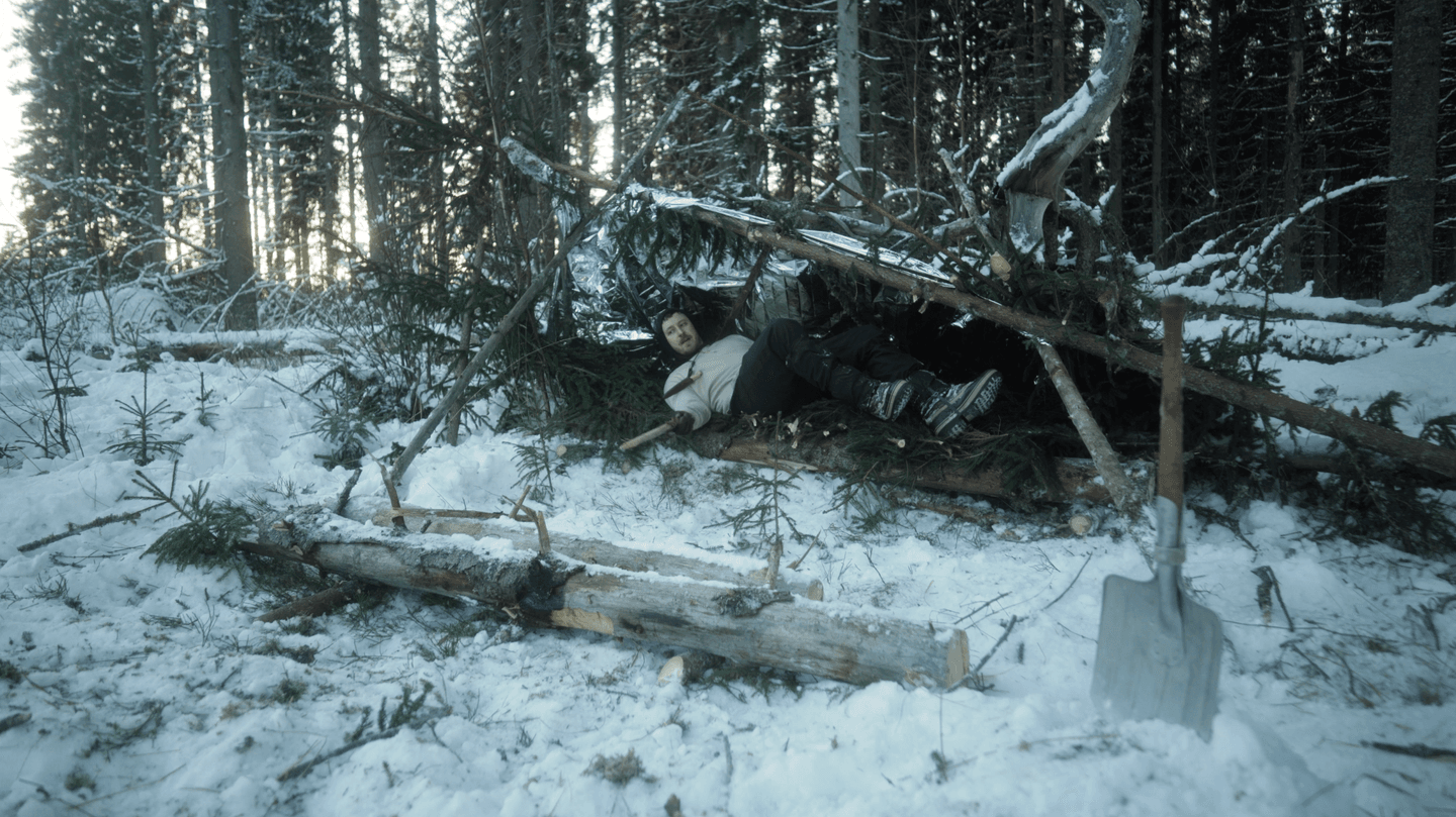 The person is lying inside a shelter made of branches and spruce twigs in winter. In the foreground, there is a shovel and two short logs side by side.