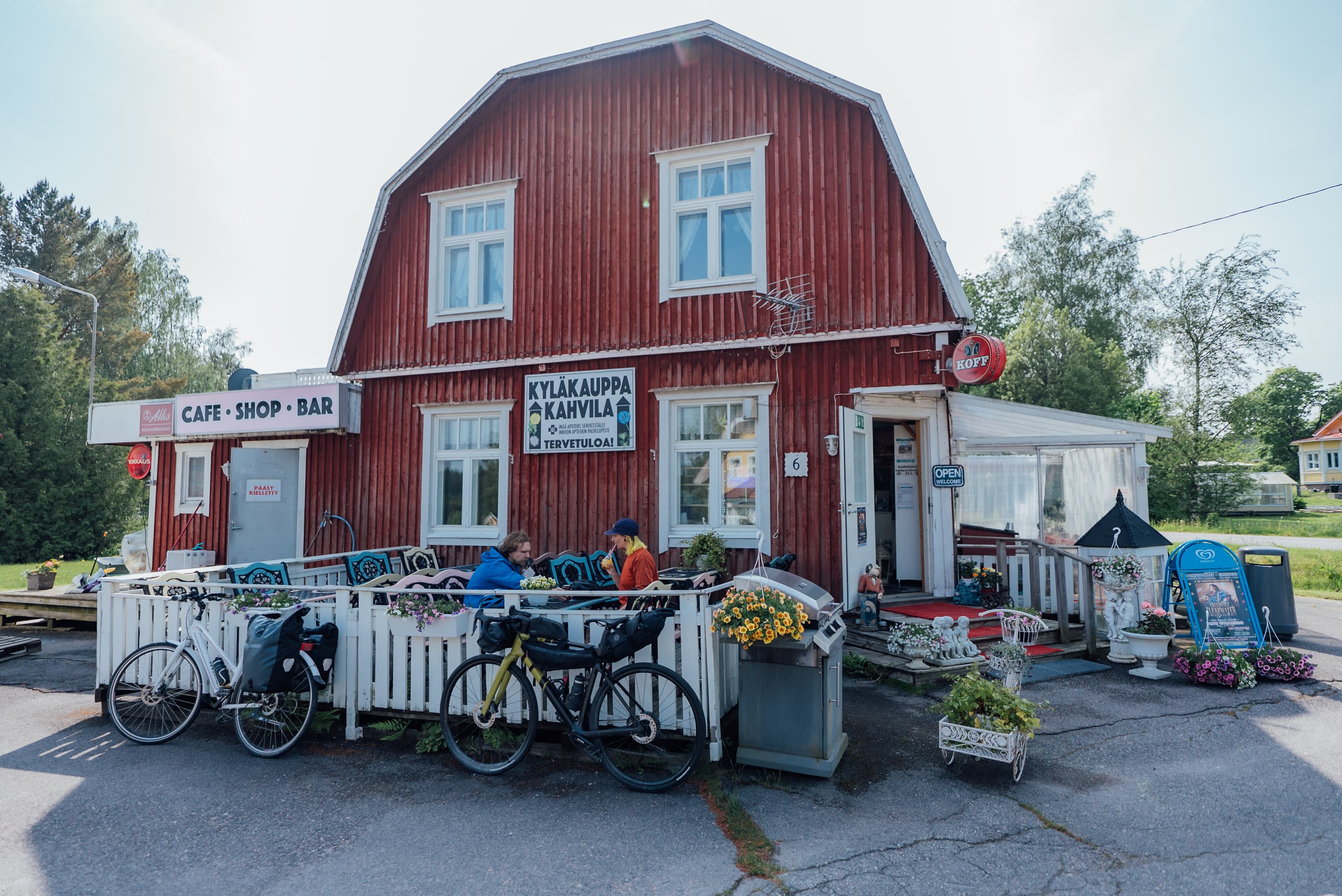 Kaksi pyörämatkailijaa viettämässä taukoa idyllisen kyläkauppakahvilan terassilla, pyörien nojatessa terassin aitaan