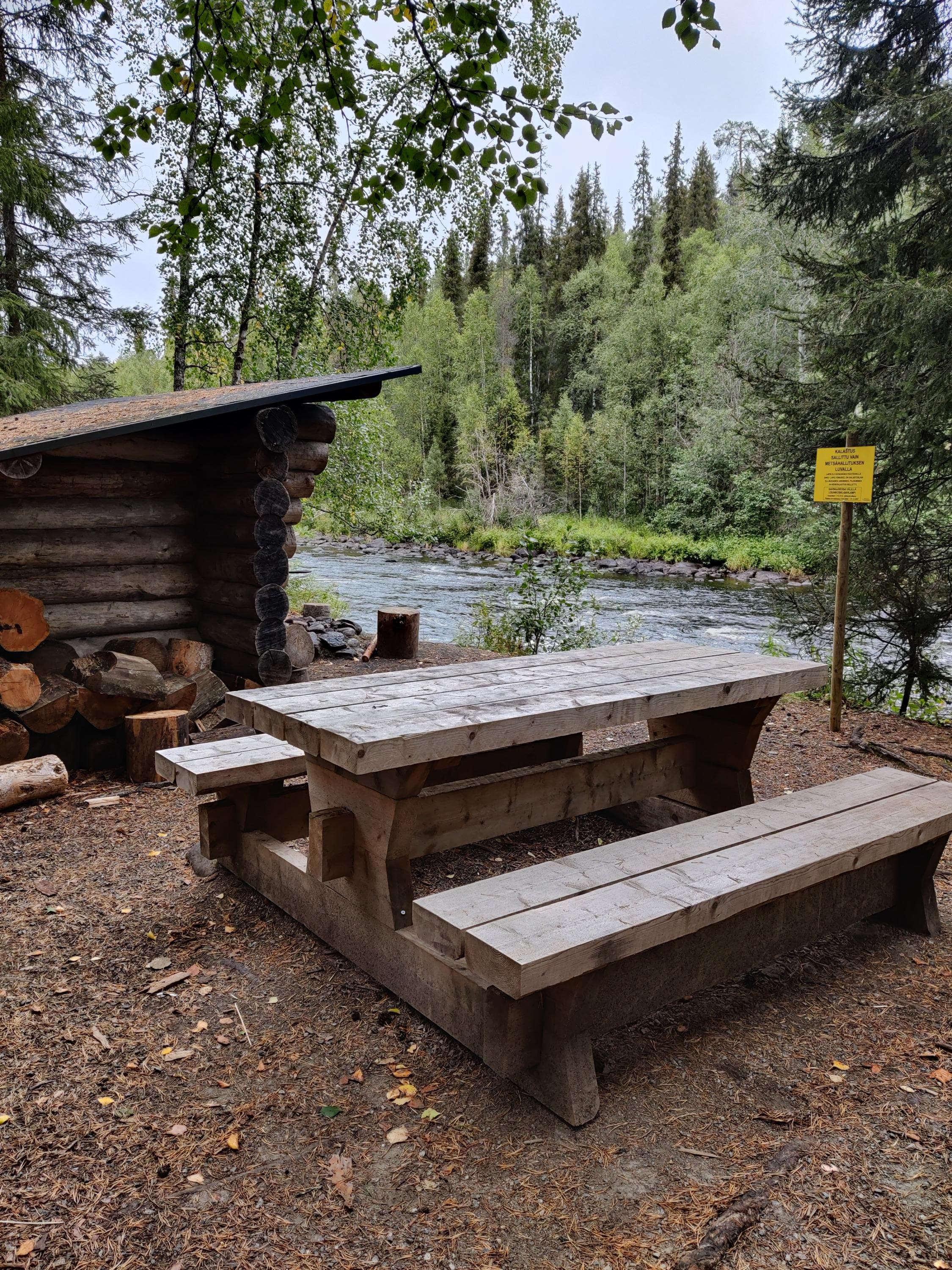 A lean-to shelter located by the river. Next to the shelter is a wooden table set.