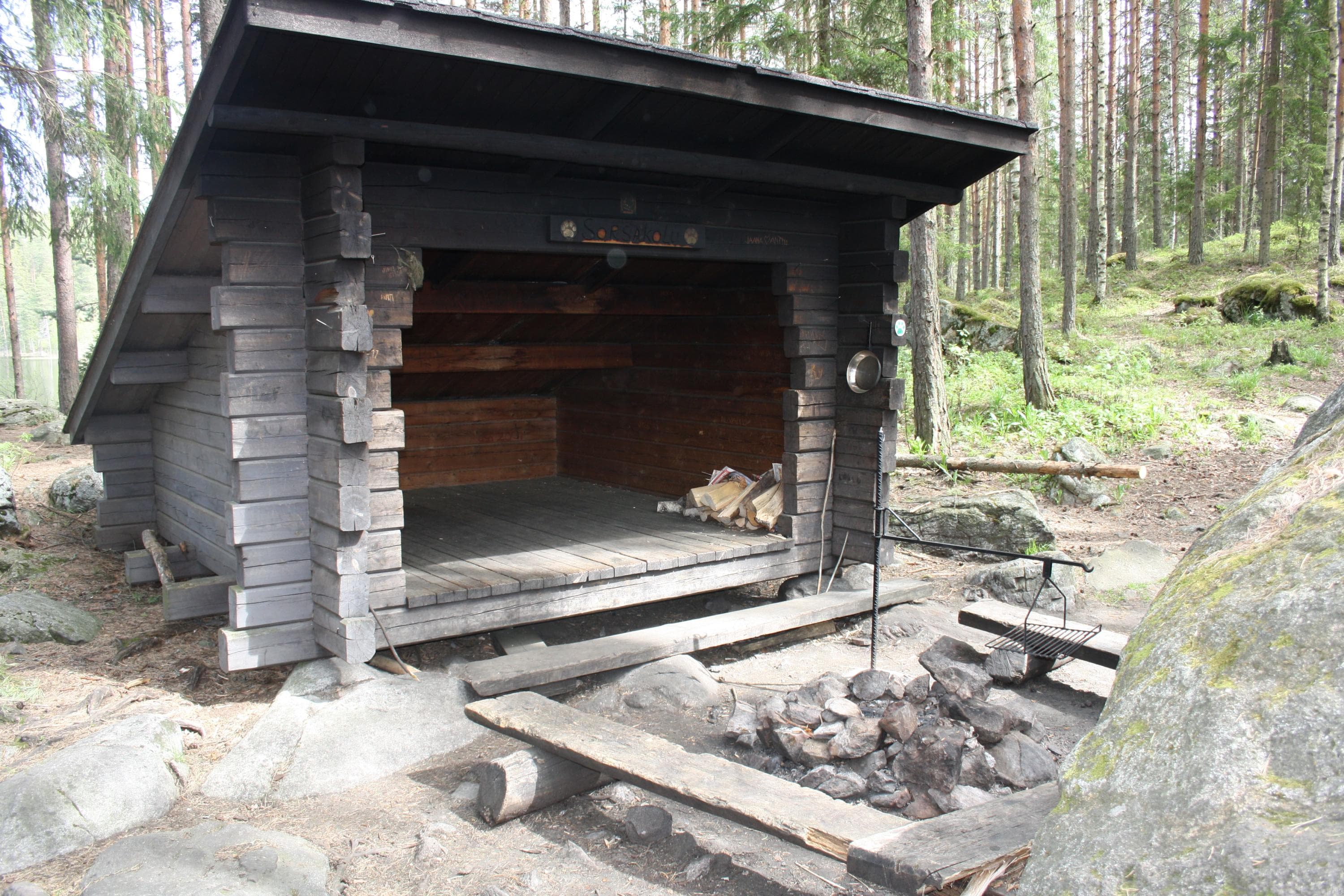 A small wooden shelter in the forest with a fire pit in front, bordered by stones.