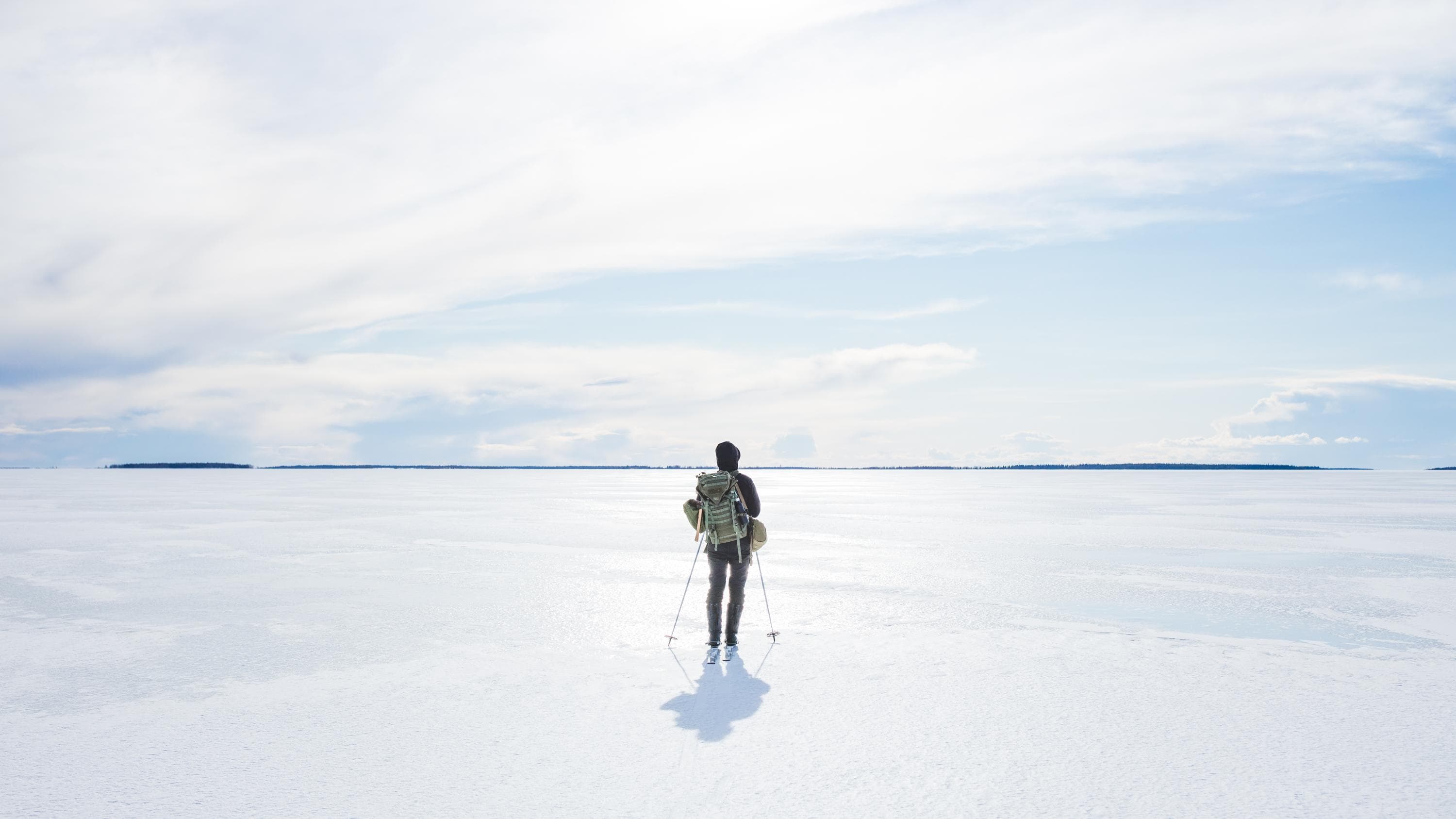 Hiihtäjä rinkkansa kanssa valkoisella jäällä. Aurinko paistaa kirkkaasti pilviverhon takaa. 