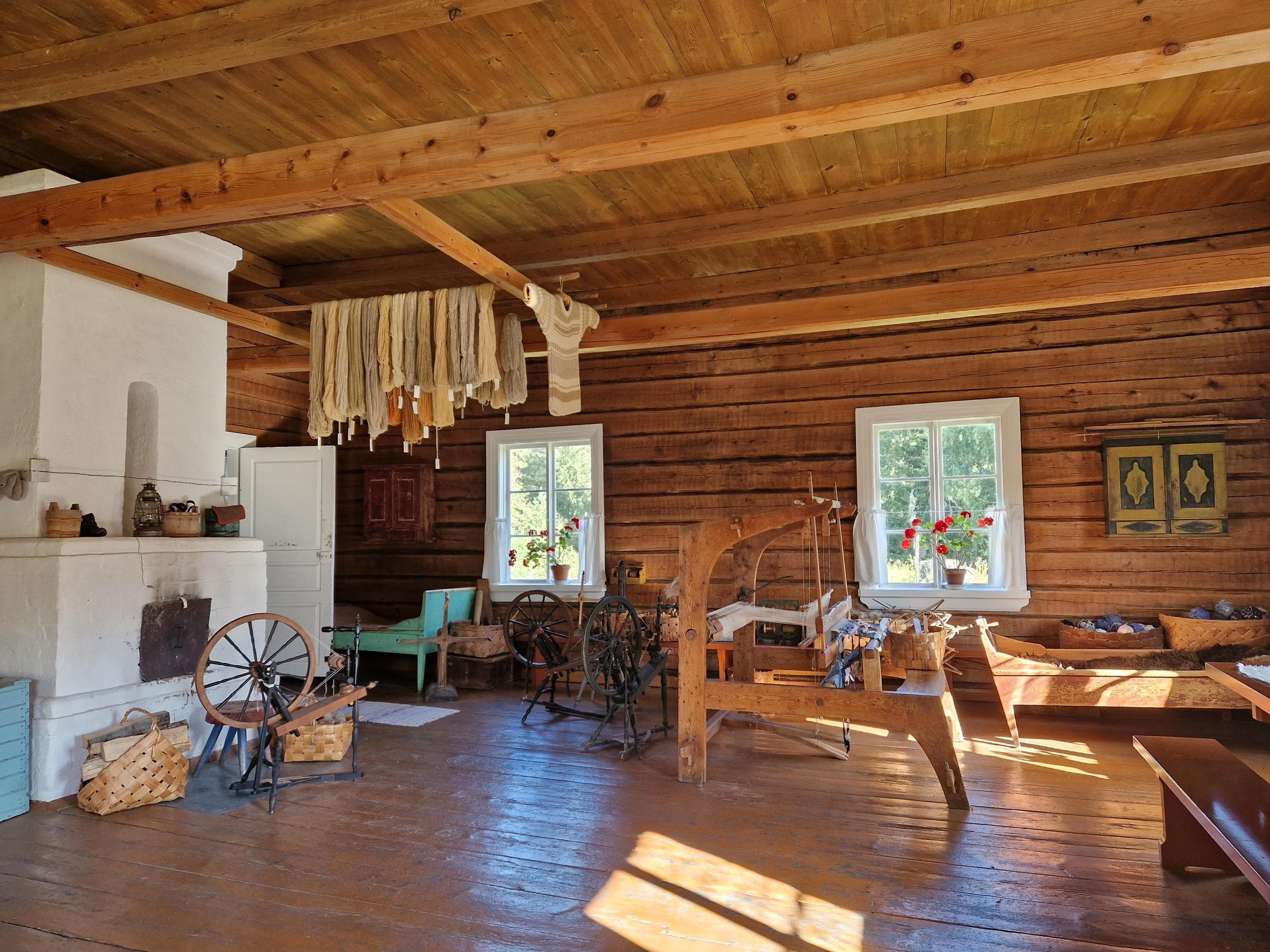 Hirsirakennuksen sisätilat. Huoneessa on suuri takka, värjättyjä lankoja ja kangaspuut.