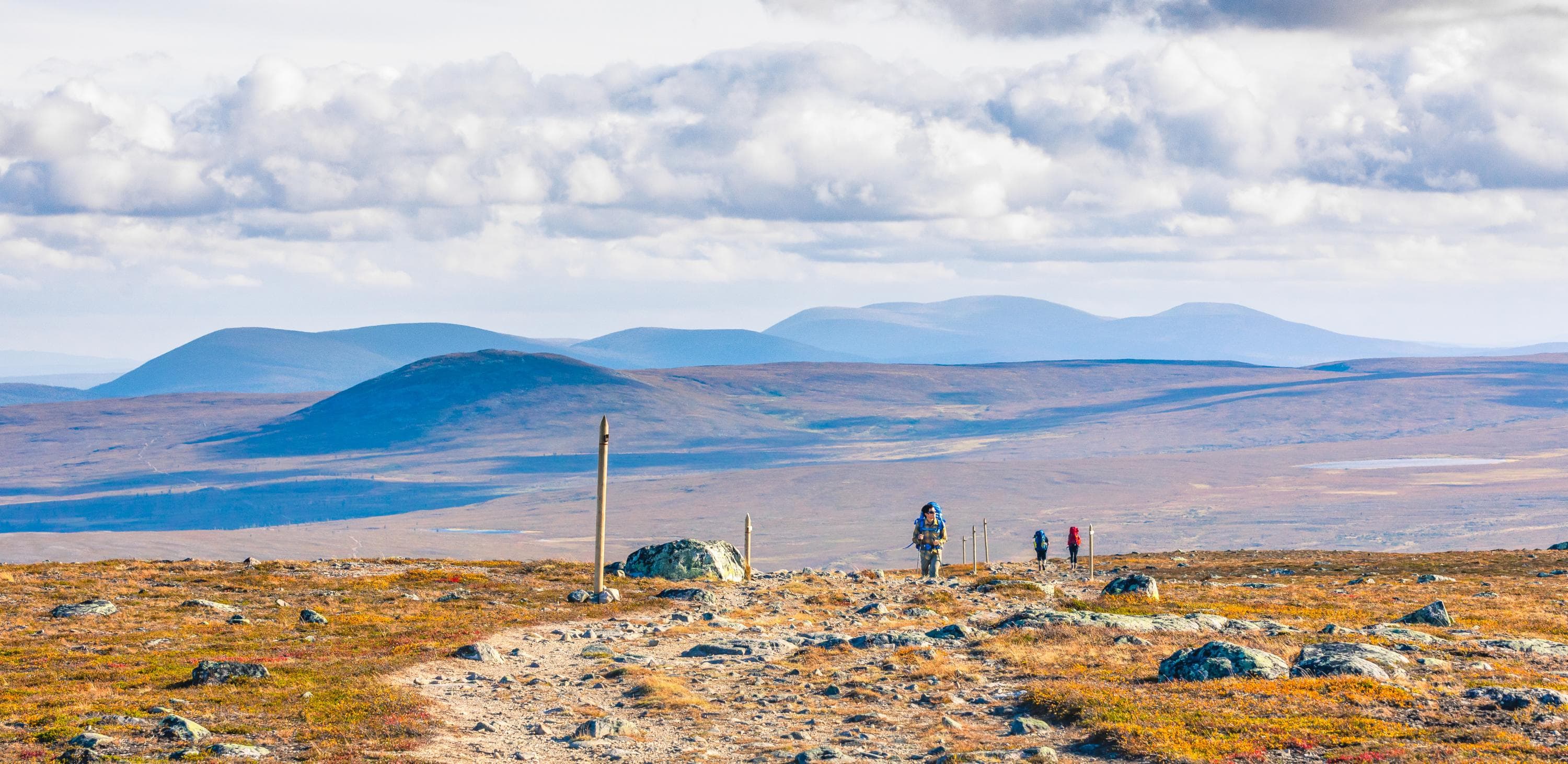 Image: Rami Valonen / Metsähallitus Three hikers on a mountain trail. In the background, a stunning row of fells.
