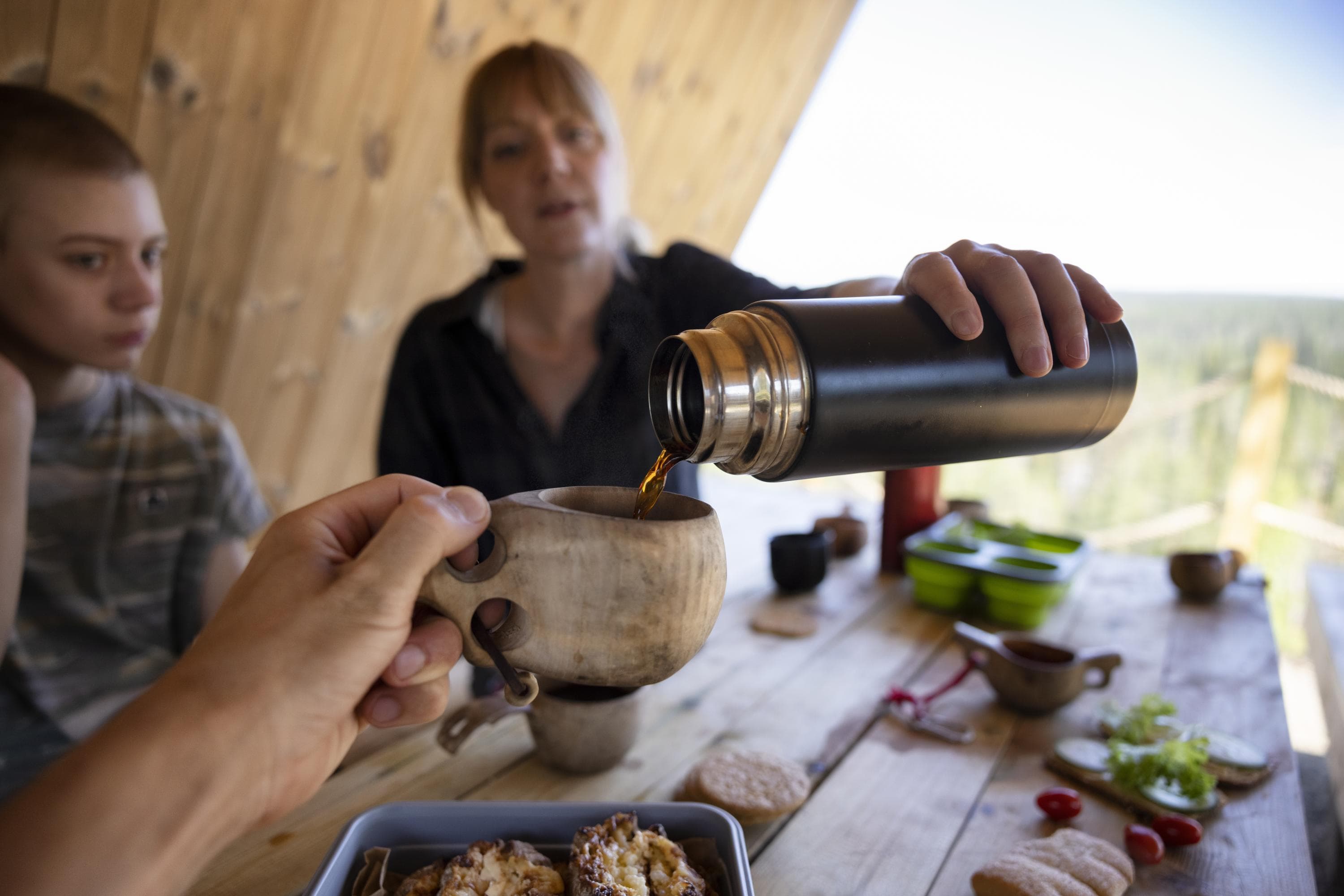 Image: Metsähallitus/Harri Tarvainen In the foreground of the picture, a hand is seen offering a kuksa. A woman is pouring coffee into the kuksa. There are snacks on the table. A child is at the edge of the picture.