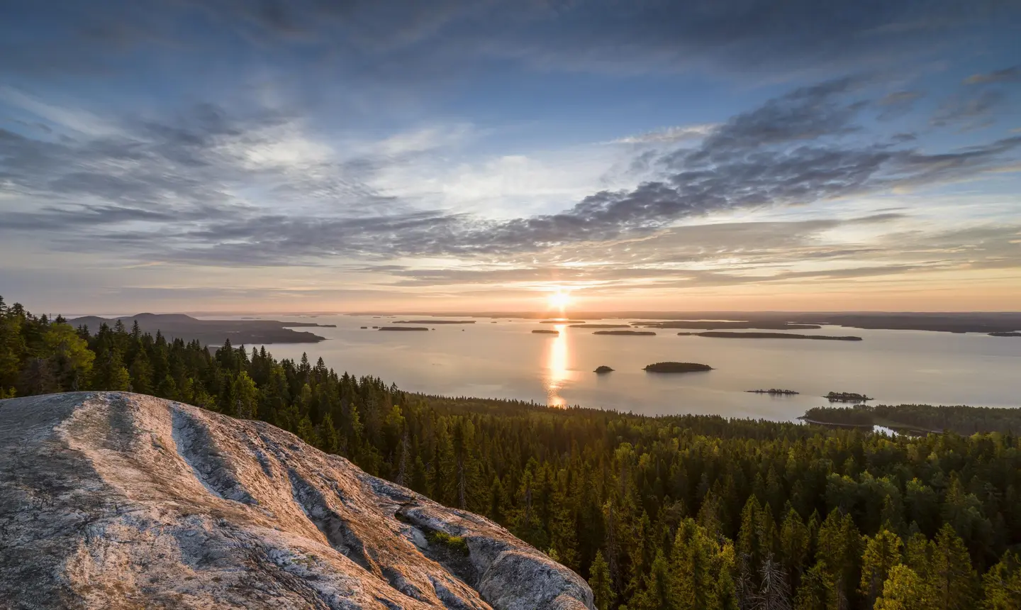 Image: Marko Haapalehto In the foreground, there is a rock and forest, with a lake view in the background. The sun is rising.