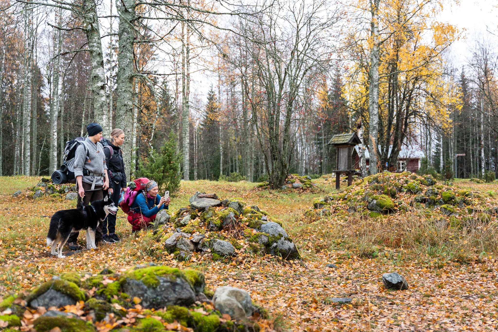 Bild: Mitja Piipponen Tre vandrare och en hund på gården av en röd stuga. En av dem fotograferar en stenhög.