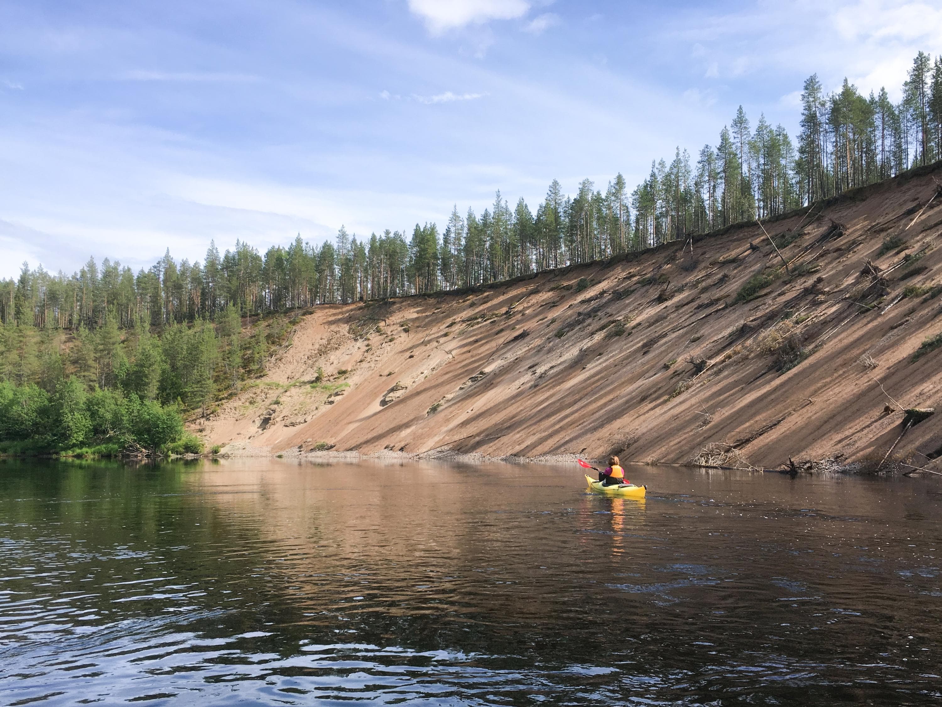 Bild: Minna Koramo En kajakpaddlare på floden. En hög sandbank kantar floden, med träd som växer ovanpå.