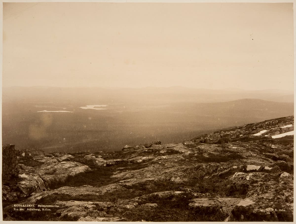 Kuva: K. E. Ståhlberg Vanha, 1800-luvun lopun mustavalkokuva kivikkoisen tunturin laelta kohti metsäistä kaukomaisemaa. Kaukana metsän keskellä siintää järvi.