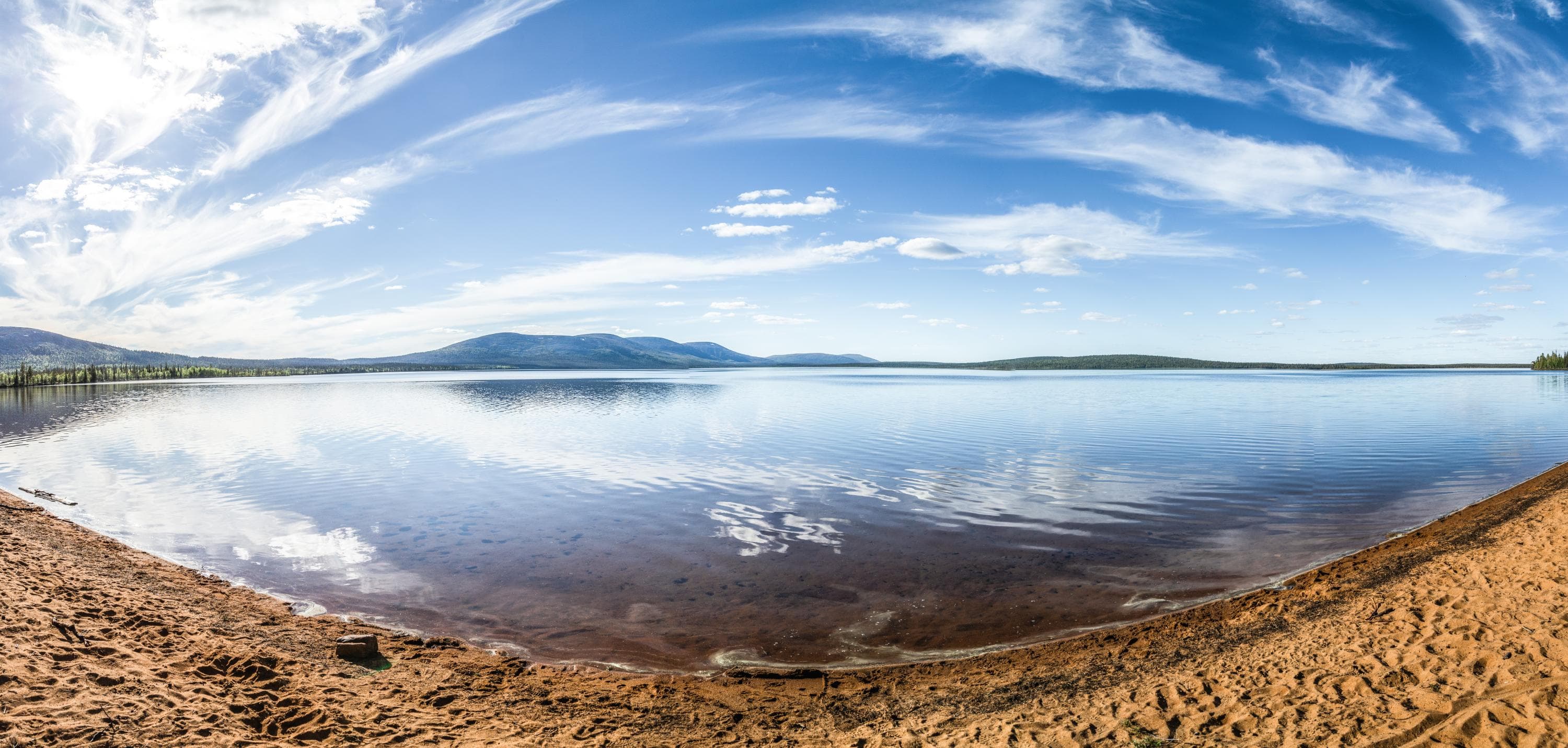 Kuva: Heikki Sulander Laajakuvassa tyyni järvenselkä, jonka takana horisontissa kapeana nauhana tuntureita. Ohutta yläpilveä sinisellä taivaalla. Etualalla hieno kivetön hiekkaranta.