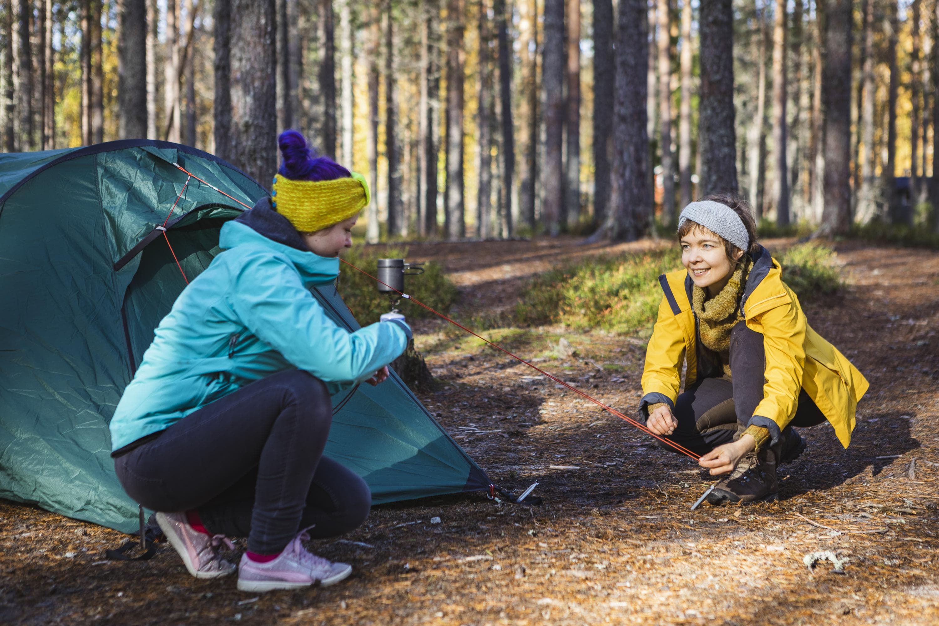 Kuva: Satu Naumanen Kaksi telttailijaa on leiritymässä ja kiinnittää teltan tukinaruja telttakiiloilla paikoilleen. Taustalla on retkikeitin kannon päässä.