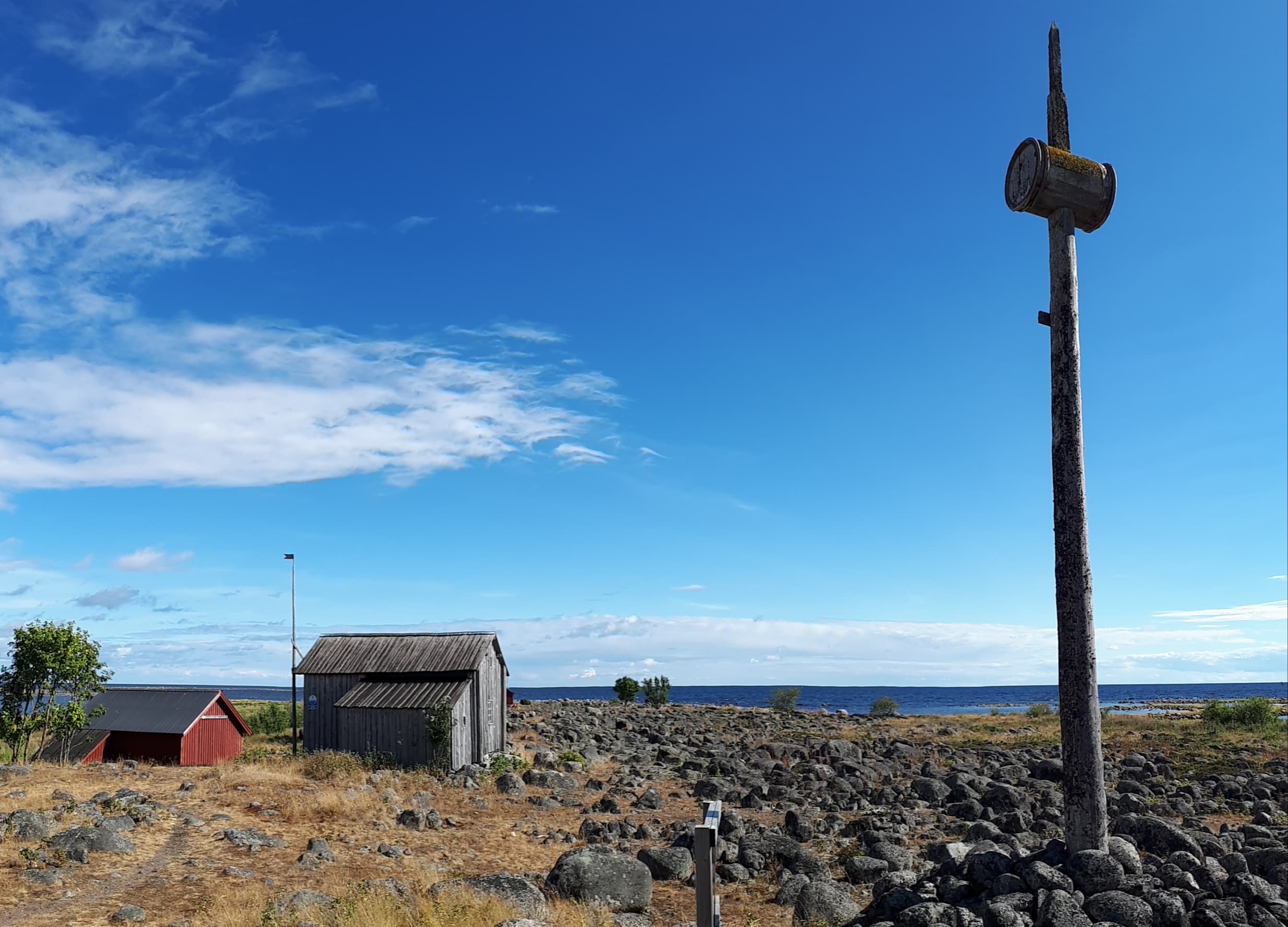 Bild: Heli Suurkuukka Två små träbyggnader bredvid ett stenblock av stora stenar. I bakgrunden syns havet och till vänster en mast med en trätunna i spetsen.