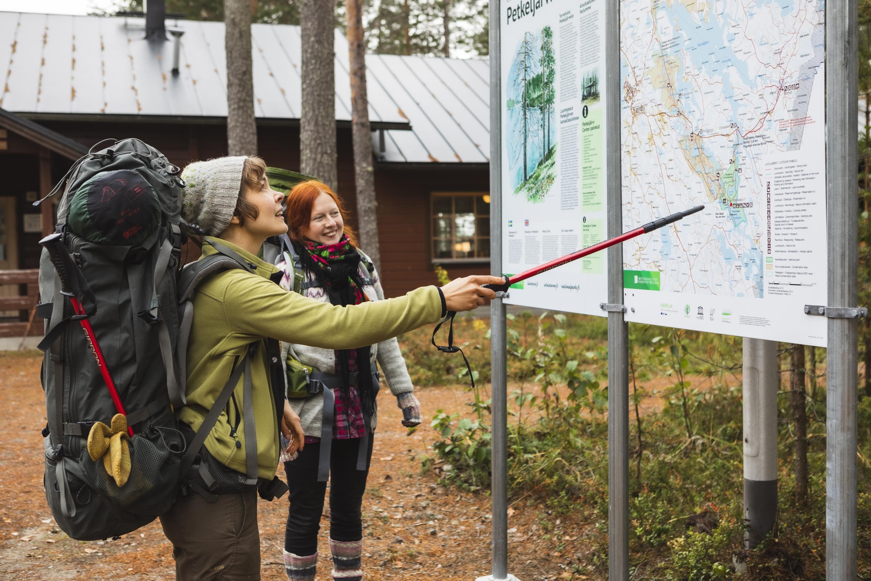 Kuva: Ulla Keituri Kaksi rinkkaselkäistä vaeltajaa seisoo tutkimassa suurta opastekarttaa. Taustalla häämöttää Patvinsuon retkeilykeskus.