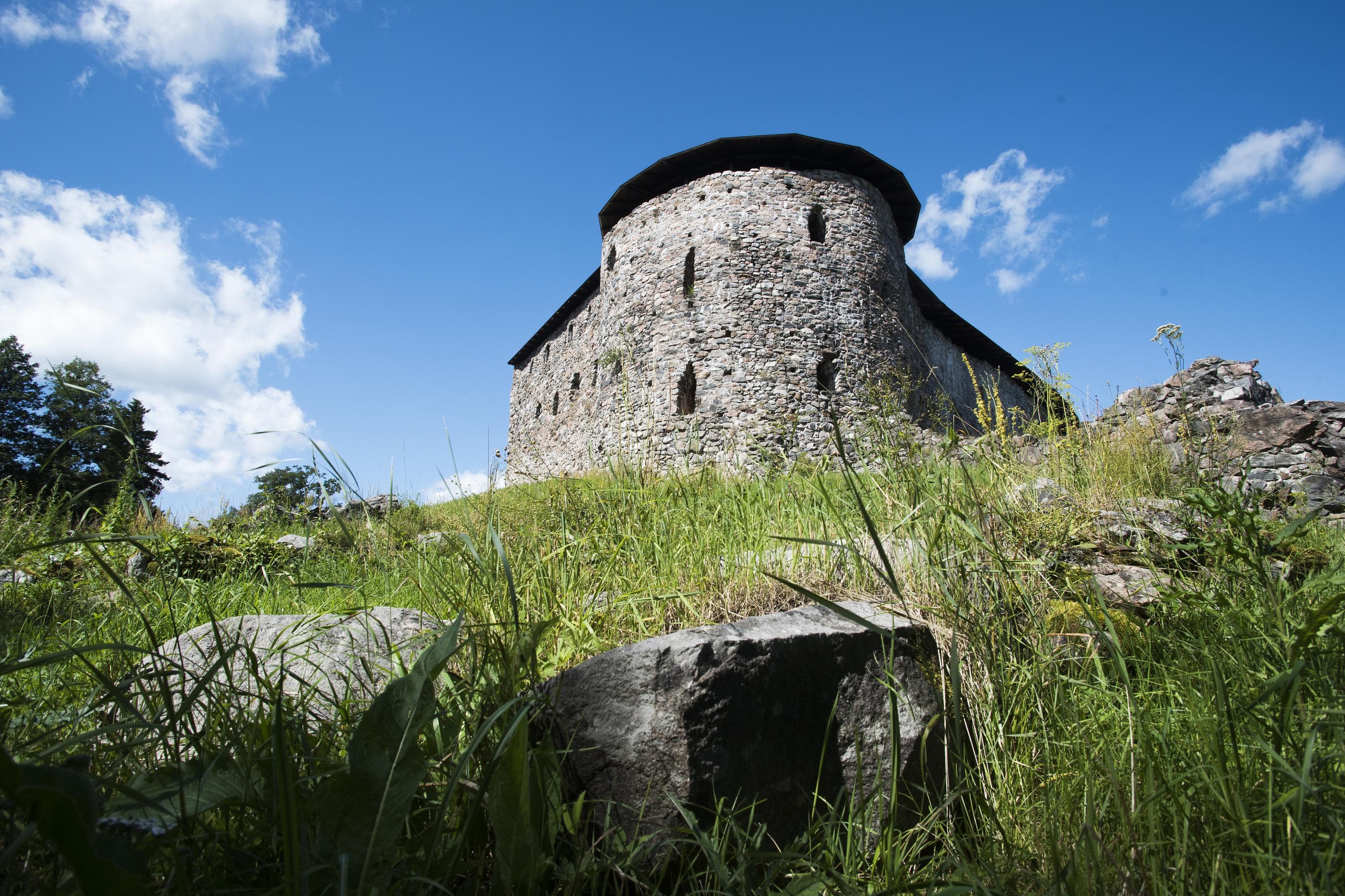 Bild: Jussi Helimäki Ruinerna av en stenborg. I förgrunden finns en stenmur och grön undervegetation. Det är moln på himlen; det är sommar.